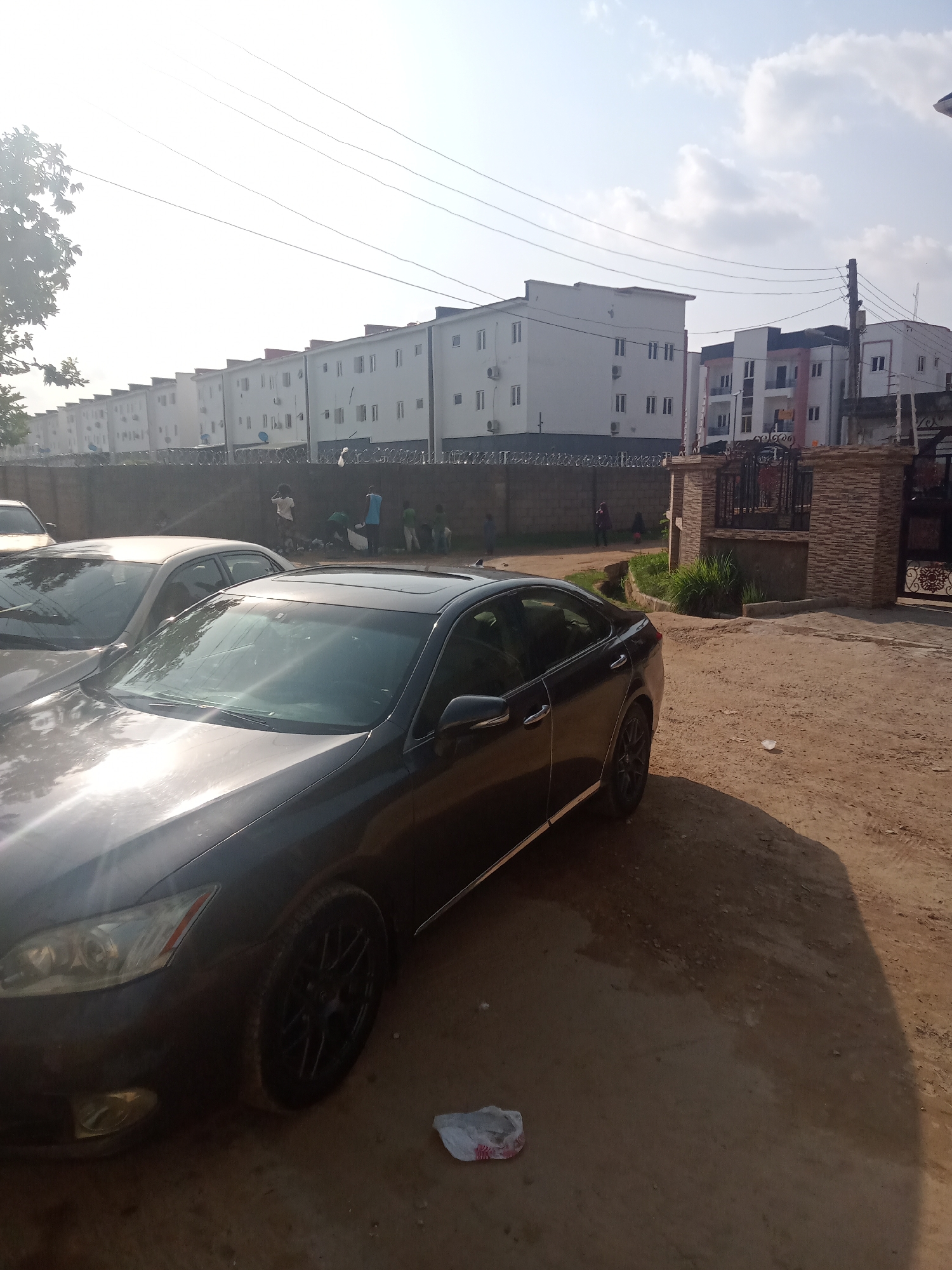The image depicts a street scene in Gwarinpa, Nigeria. In the foreground, a dark sedan car is parked on a dirt road. Another car is visible parked next to it. Behind the cars, a long row of white buildings stretches into the distance, topped with red roofing. A brick wall topped with barbed wire runs along the perimeter of the buildings.
Several individuals appear to be standing near the wall, engaged in activities like cleaning. To the right, there is a gated entrance with brick pillars, a black wrought-iron gate, and some green shrubbery. Overhead, power lines stretch across the sky. The sky is light blue with a few clouds.