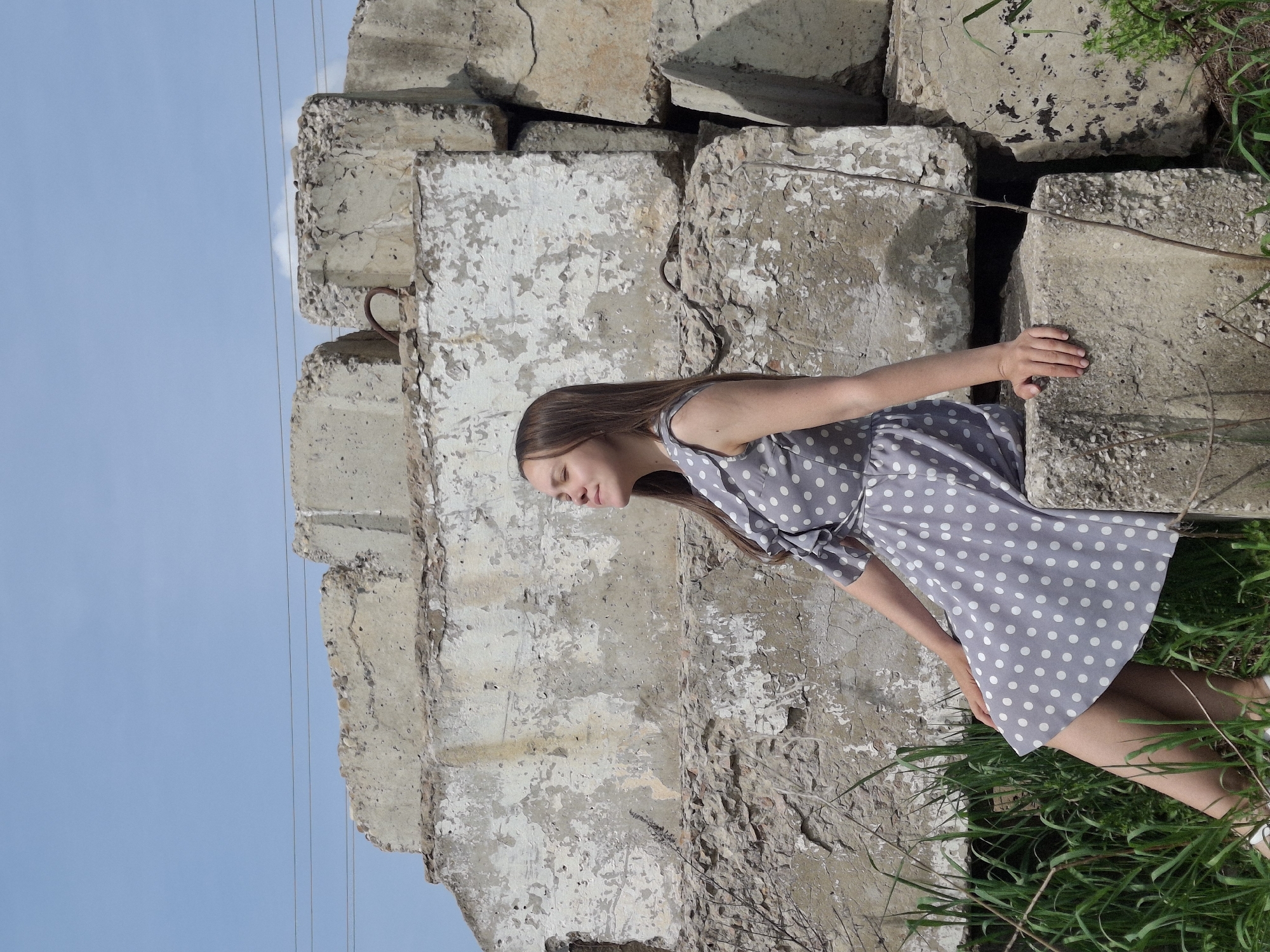 A young woman in a gray and white polka dot dress is leaning against a stack of weathered, concrete blocks in what appears to be an outdoor location in the city of Chaikovsky, Russia. The blocks are stacked haphazardly and show signs of age, with peeling paint and exposed aggregate. The woman's eyes are closed and her expression is serene. She is positioned between the concrete blocks and some green grass and vegetation. The background features a blue sky, and thin electrical wires are visible near the top left of the frame.