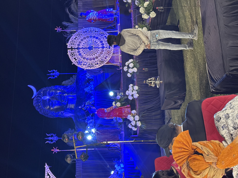 A scene is captured at night with a large, blue-lit statue of Shiva, adorned with a crescent moon. A person in a light jacket and jeans stands in front, looking at the statue. Floral arrangements and decorative lights enhance the ambiance. In the foreground, attendees dressed in traditional clothing are seated, their backs to the camera. An ornate white circular decoration hangs above the statue.