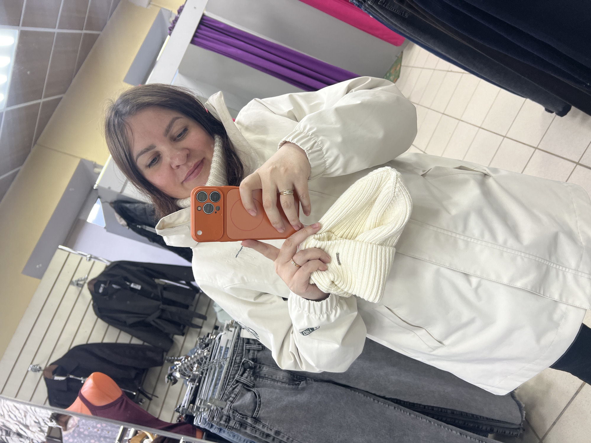 A young woman is captured in a mirror selfie within what appears to be a clothing store or changing room in Perm, Russia. She is positioned slightly off-center, holding an orange smartphone with a multi-lens camera in her right hand, aiming it towards the mirror to take the photo. A simple band-style ring is visible on her right ring finger.
She has brown hair and is wearing a light-colored (cream or off-white) high-neck sweater or turtleneck, covered by a matching light-colored, long-sleeved jacket or coat. In her left hand, she holds a knitted, cream-colored beanie or hat, which has a small, dark rectangular label visible on it. Her expression is gentle and focused, as she looks down at her phone screen. The attire suggests cooler weather, possibly autumn or winter.
The background indicates an indoor retail space. Clothing racks with various garments, including dark jackets and jeans, are visible behind and to the sides of the woman. A purple curtain, commonly found in changing rooms, is reflected in the upper left of the mirror. The floor is tiled with light-colored square tiles. Overhead, part of a white grid ceiling with fluorescent lighting is also reflected. Some white text, partially reversed due to the mirror and obscured, is visible on the sleeve of her jacket. The scene depicts a casual shopping moment, likely while trying on or considering new items.