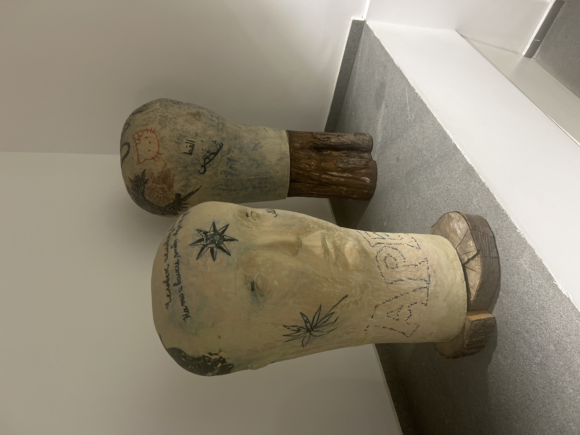 Two sculptural head busts are displayed side-by-side in what appears to be an indoor exhibition space with white walls and a grey floor. The busts are made of a light-colored material, possibly plaster or painted wood, and are mounted on rough, dark wooden bases that resemble tree stumps or logs.
The closest bust depicts a human face in a neutral expression and is extensively decorated with drawings and text. On its left side, Russian text reads "Человек Человеку" (Man to Man) followed by "На то и волки, чтобы..." (That's what wolves are for, to...). A prominent black star-like symbol or compass rose is drawn on the forehead, and a stylized black leaf (resembling a cannabis leaf) is visible on the cheek. Other black, abstract patterns and symbols adorn the lower part of the bust.
The second bust, positioned slightly behind the first, is also decorated. A distinct red outline drawing of Hello Kitty is visible on its upper side. Below this, Arabic script is written in black, with "شخص" (Shakhs, meaning 'Person') and "القط" (Al-Qitt, meaning 'The Cat') clearly discernible. Other dark, less defined markings are present around its surface.
Both sculptures rest on their rustic wooden bases on the grey floor, against the plain white wall of the setting. The lighting is even, suggesting an indoor, controlled environment, possibly a gallery or a modern public space in Sofia, Bulgaria, given the context. The sculptures appear to be contemporary art pieces combining different cultural references and artistic styles.