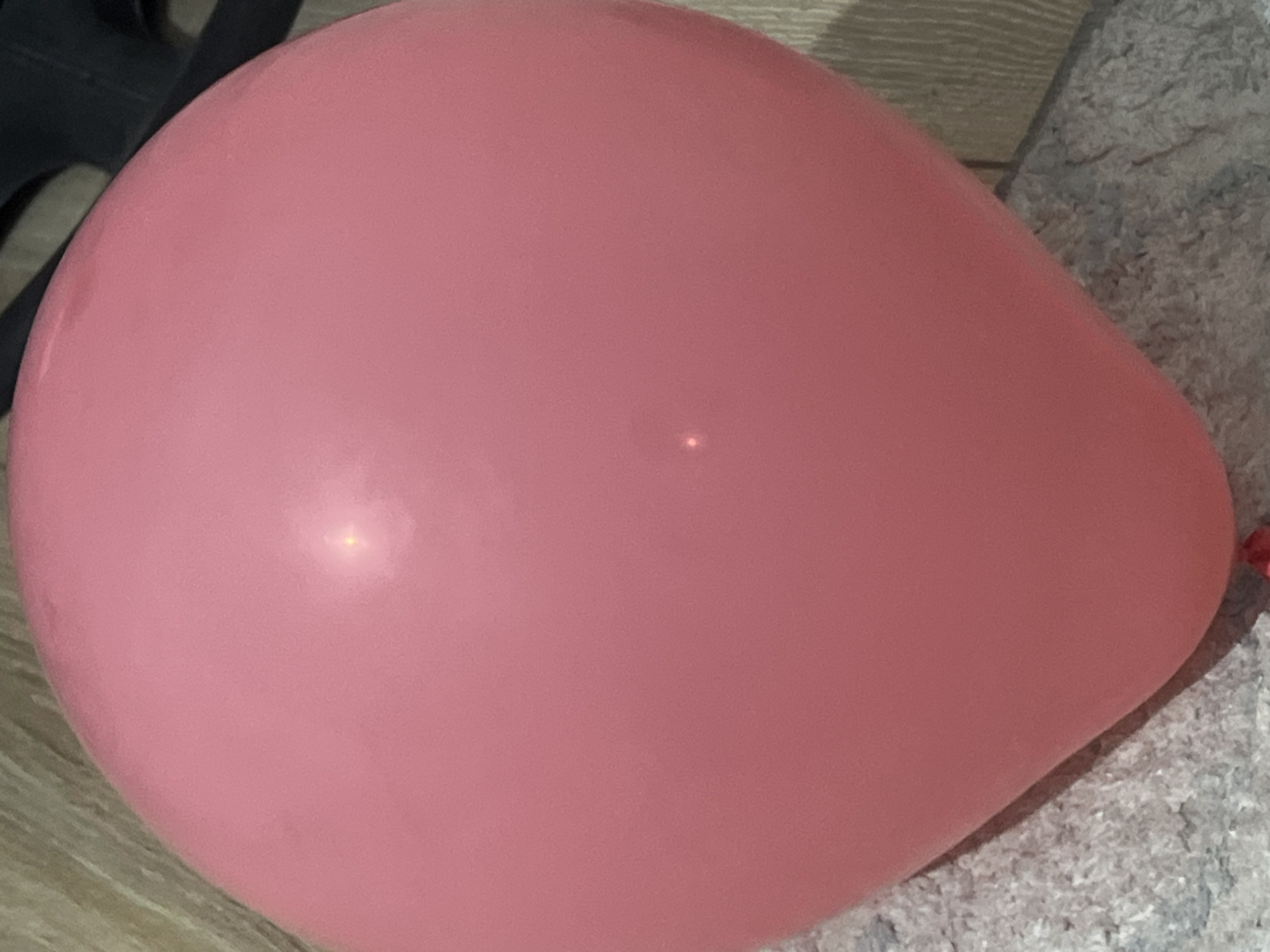 A close-up, eye-level, vertical shot features a large, pink, inflated balloon. The balloon fills most of the frame, its surface smooth and reflecting light in several small, bright spots. The bottom of the balloon rests on a textured surface that appears to be a light-colored, possibly grey, carpet. Behind the balloon, to the right, a vertical wooden-patterned surface is visible, suggesting a wall or furniture. The lighting is somewhat dim, with a strong highlight on the balloon's surface. The scene is devoid of any people or overt activity.