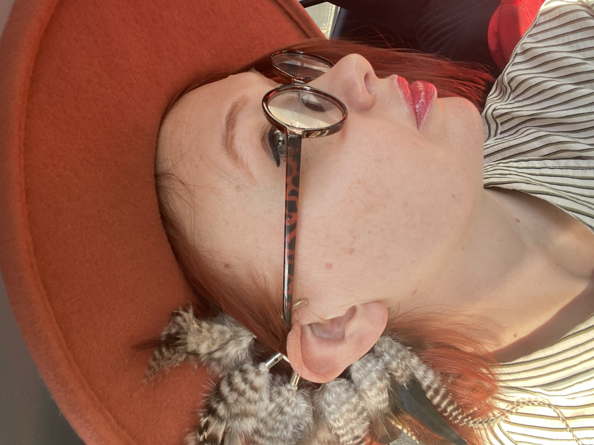 This is a close-up, rotated portrait of a person, likely taken in Yekaterinburg, Russia. The individual is seen in profile, looking upwards or to their right. They have fair skin, with visible freckles or moles, and reddish-brown hair peeking from under an orange or rust-colored felt hat, which occupies the left side of the frame.
The person is wearing round glasses with tortoise-shell frames, resting on their nose. Their lips are painted with a glossy red lipstick. Prominently featured are large, ornate feather earrings, crafted from various natural feathers in shades of grey, brown, and white with distinct patterns, dangling from their right ear. The person is dressed in a light-colored shirt with thin horizontal stripes.
The scene is illuminated by bright natural light, indicating it is daytime. The background is indistinct, showing dark and lighter areas, possibly suggesting an indoor setting or a shaded outdoor location. The person appears to be posing for the photograph, with a calm or contemplative expression. No specific activity or event is taking place beyond this pose. There is no visible text in the image.