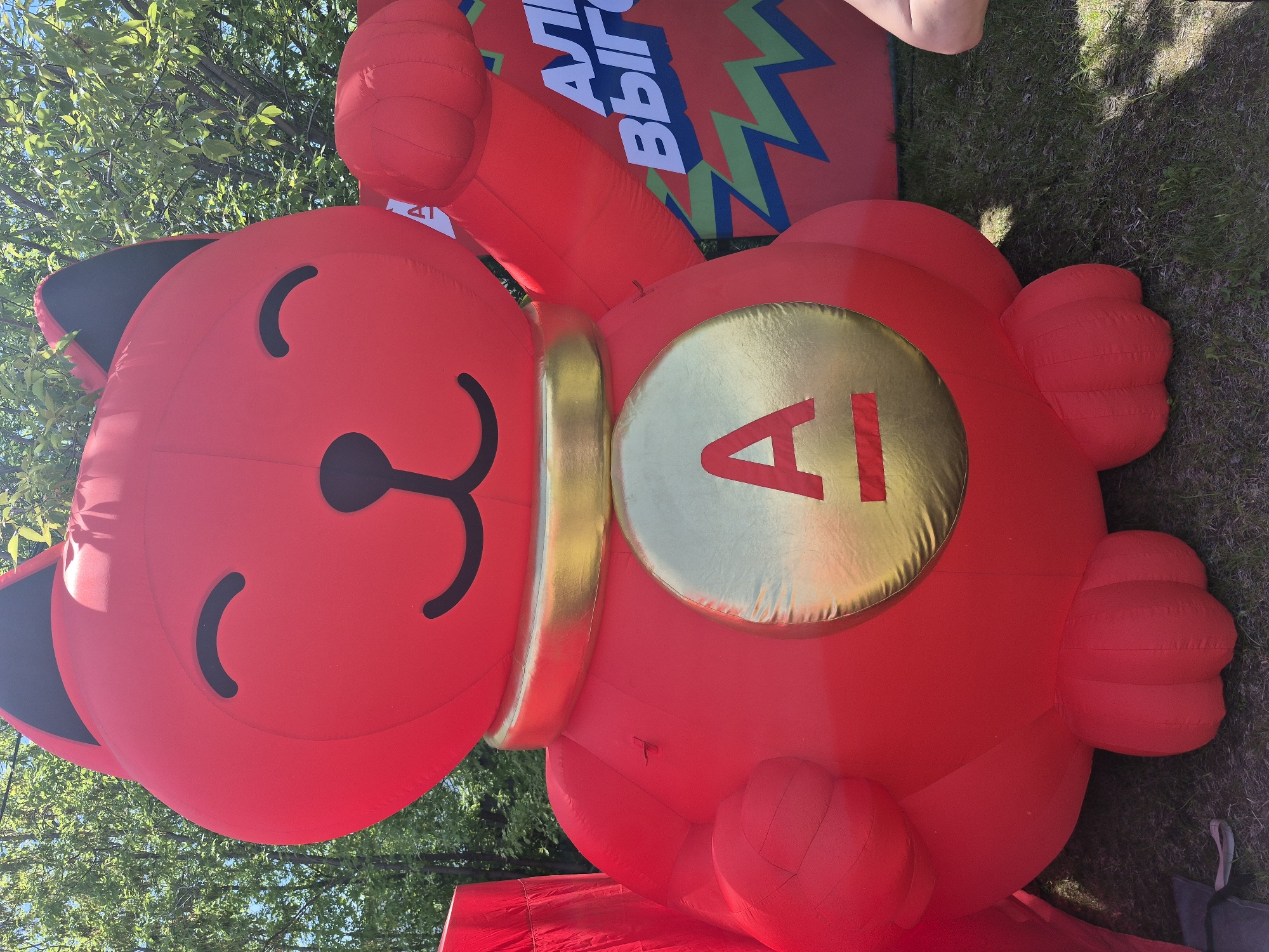 An inflatable red cat figure, styled as a Maneki-neko, is depicted outdoors in the city of Kansk, Russia. The large, red inflatable features black crescent-shaped eyes, a black triangular nose, and a black smiling mouth. Its inner ears are also black. A golden inflatable collar encircles its neck, and a prominent golden inflatable medallion with a red capital letter "A" and a horizontal red line beneath it is centered on its belly. The cat's right paw is raised, consistent with the traditional Maneki-neko gesture. Its lower body and three-lobed feet are visible, resting on green grass. In the background, lush green tree foliage is visible under a clear sky. To the right of the red cat, a section of another large, red inflatable object is partially visible. This background inflatable displays white Cyrillic text, reading "АЛЫ" and "ВЫГ", likely fragments of longer words, above zigzag patterns in green, blue, and black. A small portion of a human limb is visible on the extreme right, and a grey fabric anchor is on the grass at the bottom left.