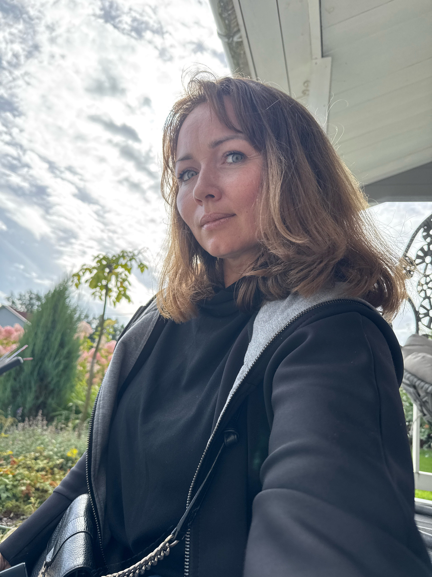 A woman with shoulder-length brown hair and blue eyes looks at the camera while outdoors. She is wearing a black hooded jacket with gray trim and a black purse with a chain strap. The background shows trees, a garden, and a cloudy sky. The image appears to have been taken in Oliki, Russia.