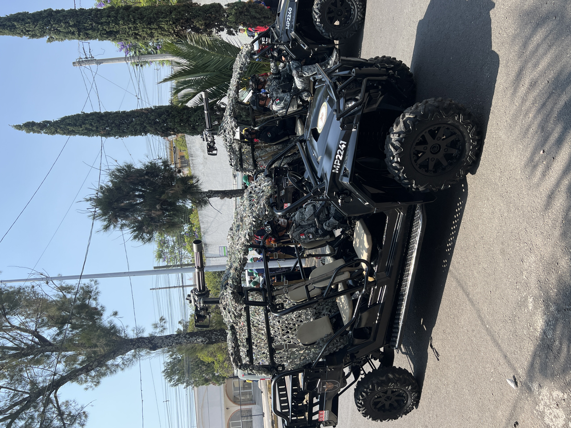 The image shows two black all-terrain vehicles (ATVs) with "MP2241" labeled on the sides, situated on a paved road in an urban environment. The ATVs are camouflaged with netting and equipped with mounted weapons. People are visible inside the vehicles, wearing military attire. Tall trees line the street and power lines are strung overhead. Buildings are visible in the background. The location is specified as an unknown city, and unknown district.