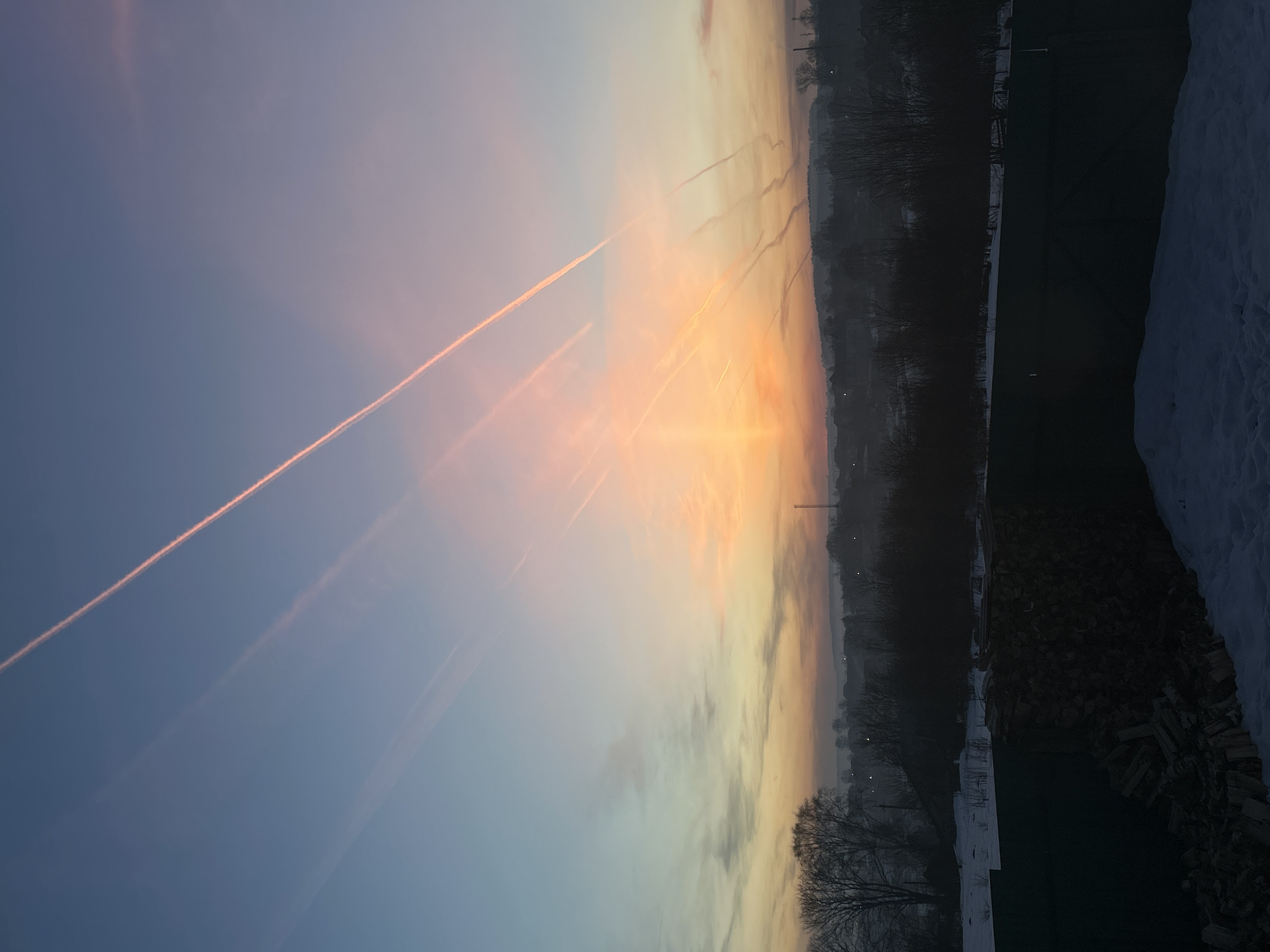 The image shows a landscape with a colorful sky at sunset. The sky transitions from a light blue to shades of orange, pink, and yellow towards the horizon. Several contrails streak across the sky, appearing as straight, faint lines. In the lower portion of the image, the ground is partially covered with snow. Trees are visible in the middle ground and appear bare, silhouetted against the sky. In the far distance, the skyline of Фоки, Russia is partially visible, marked by a tall structure resembling a crane or tower. The foreground contains a dark green area, potentially a lawn, bordered by piles of logs and more snow-covered areas.