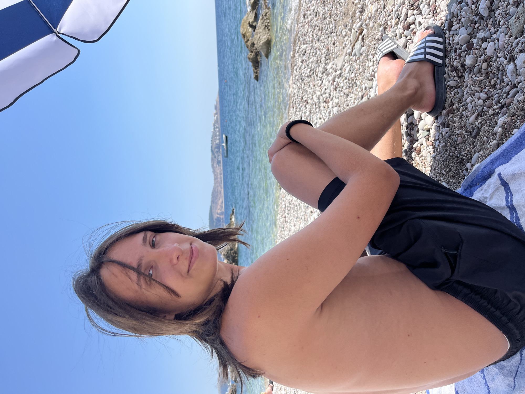 A young person with long, brown hair is sitting on a pebbled beach in Bar, Montenegro. They are wearing black shorts and black and white striped slides. The person is shirtless and is looking towards the camera with a slight smile. The sea is visible behind them, with a boat in the distance and hills on the horizon. Part of a blue and white beach umbrella is visible in the upper right corner of the frame. The sky is clear and blue. The ground is covered in small, grey and white pebbles.