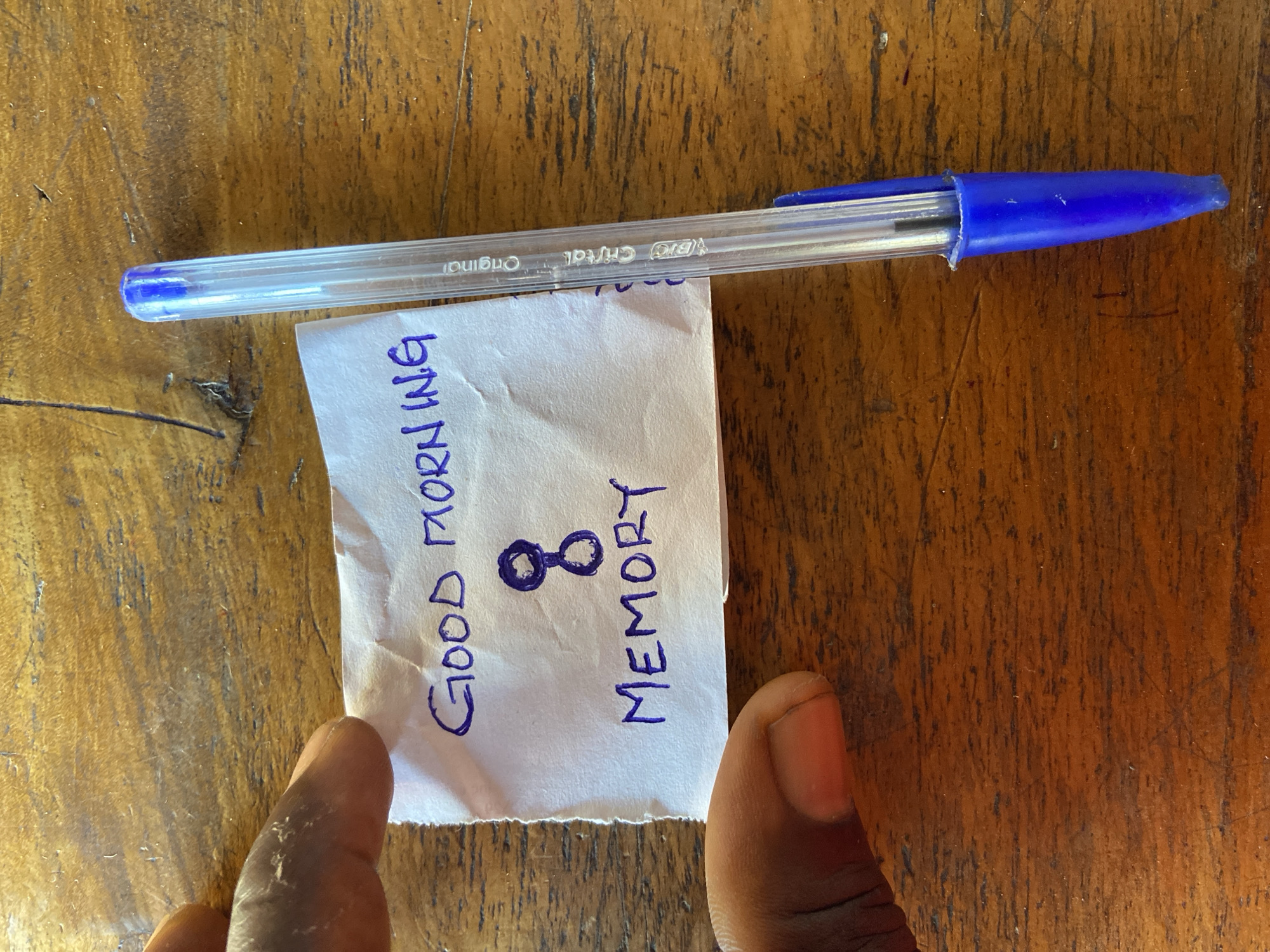 A close-up, top-down view captures a crumpled piece of white paper on a wooden surface. The paper displays handwritten text in blue ink. The top line reads "GOOD MORNING" and below it is a stylized drawing resembling an hourglass or infinity symbol. Underneath the symbol, the word "MEMORY" is written. To the right of the paper, a clear blue Bic Cristal pen lies vertically, its tip pointing downwards. A person's thumb and index finger are visible on the left side of the paper, holding it down. The wooden surface appears to be a table, with a visible grain pattern and some imperfections. The scene is set in Mararaba, Nigeria.