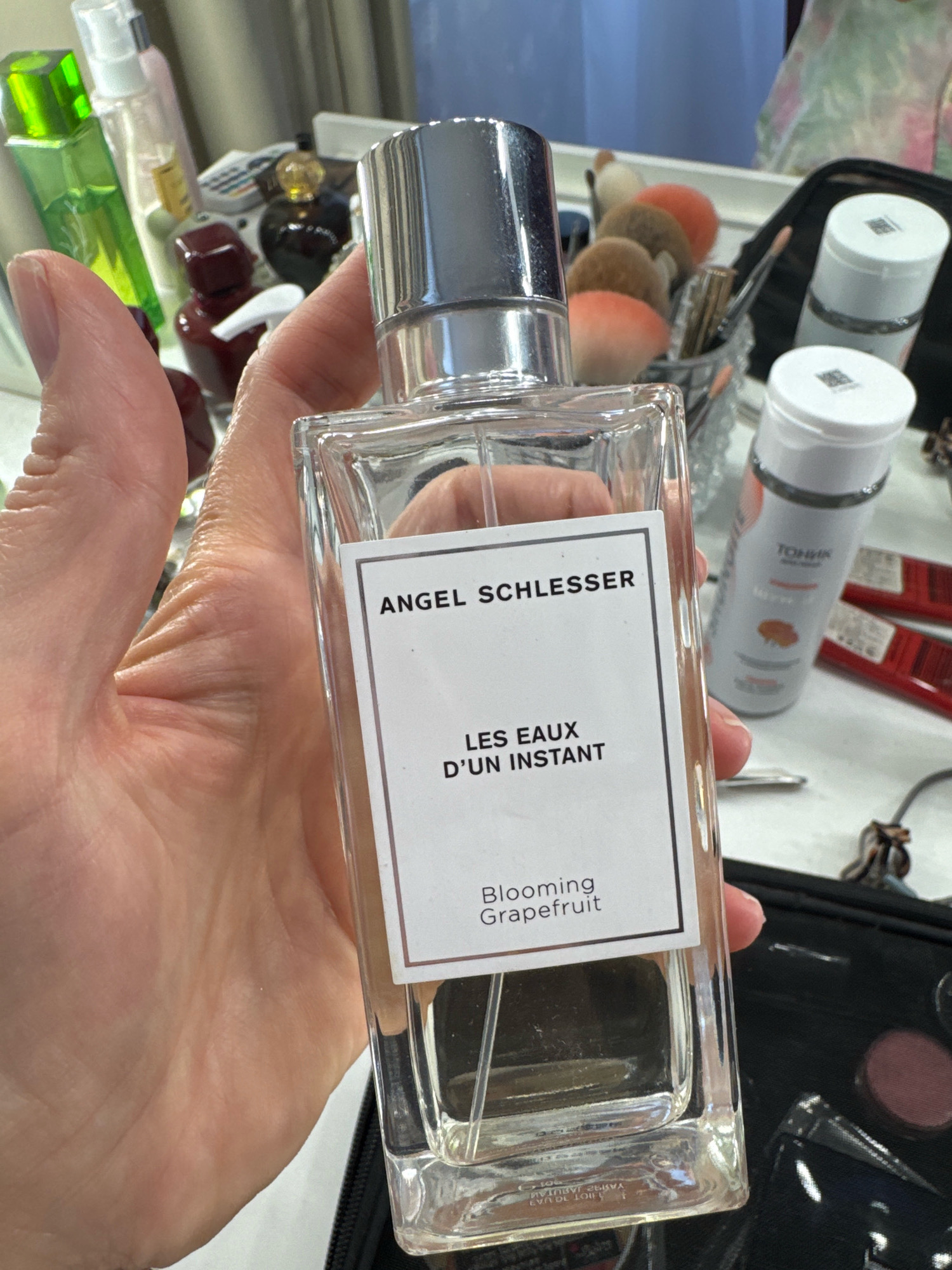 A person's hand is holding a clear glass bottle of Angel Schlesser perfume with a silver cap. The label on the bottle reads "ANGEL SCHLESSER LES EAUX D'UN INSTANT Blooming Grapefruit". In the blurred background, there are several other bottles, makeup brushes, and cosmetic items. The scene appears to be indoors, possibly a dressing table or vanity area. There is no indication of the city of Ladozhskoe Ozero, Russia, in the image.