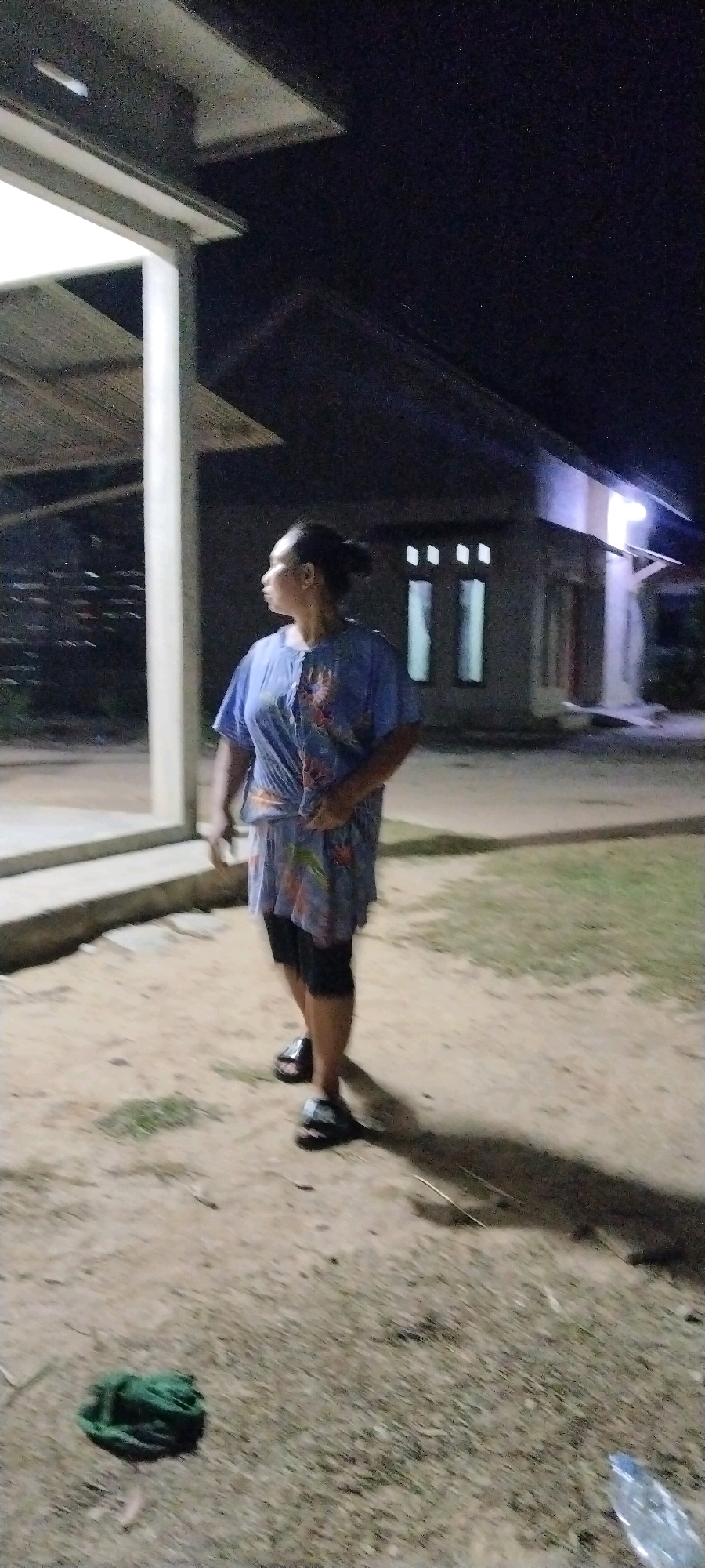 An Indonesian woman with dark hair pulled back stands outdoors at night, facing to her left, with her head turned to look in that direction. She is wearing a blue, short-sleeved, patterned shirt and dark shorts. She has on dark sandals. The ground is sandy and dirt, with some dry grass and twigs. To her left is a white pillar and a section of a roof with a textured underside, extending from a building. In the background, there is a building with several illuminated windows, and the area is dark. A green coiled object is on the ground in the lower left portion of the image, and a plastic water bottle is visible in the lower right. The image appears to have been taken in the area of Tanjung Ratu Ilir, Indonesia.