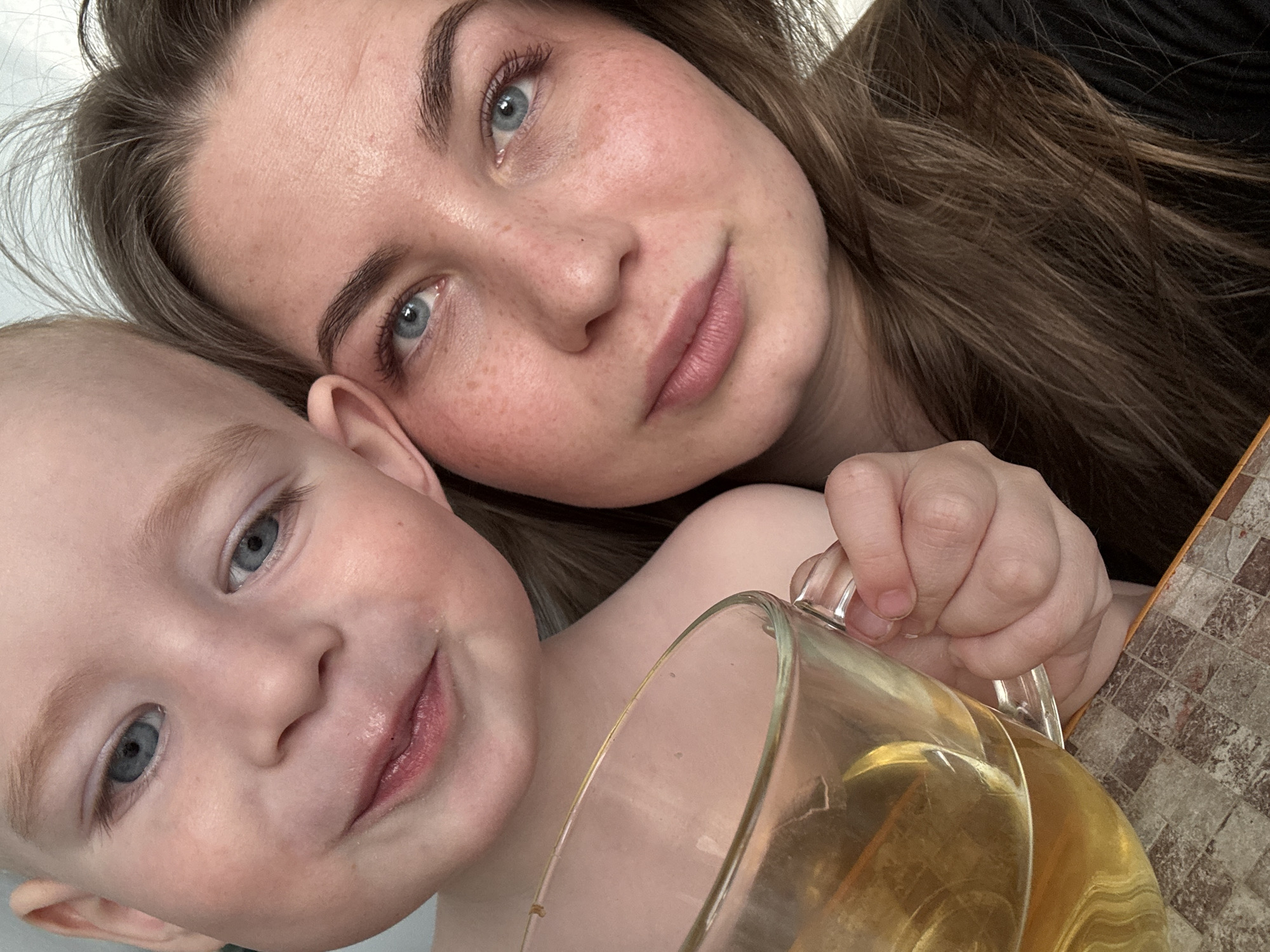 A close-up selfie shows a fair-skinned woman and a young child, likely a toddler, both with blue eyes and light hair. The woman's face is close to the camera, positioned above and slightly to the right of the child. She has freckles and her hair is brown with light highlights. The child is located on the left side of the frame. A glass mug containing a yellow liquid is partially visible in the lower right foreground, held by the woman's hand. A patterned surface is also visible in the background, suggesting a table. The location is likely a home in the city of Chaikovsky, Russia. The interaction suggests an intimate moment between a parent and child, potentially during a beverage break.
