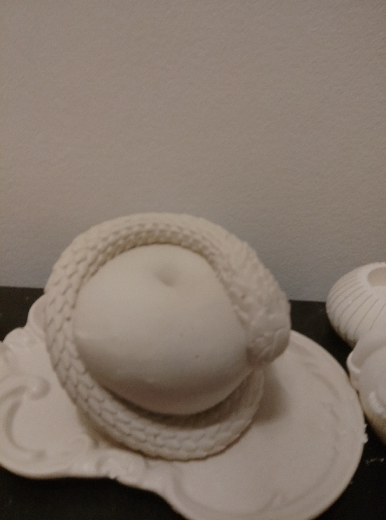 This image depicts a close-up, unedited view of a white ceramic sculpture resembling a snake coiled around a ball, set against a neutral background. The sculpture is placed on a decorative white ceramic plate. The lighting is somewhat dim, casting subtle shadows. No people are present, and no location in Wedel, Germany, is visible or implied. The image is focused on the texture and form of the sculpture itself.
