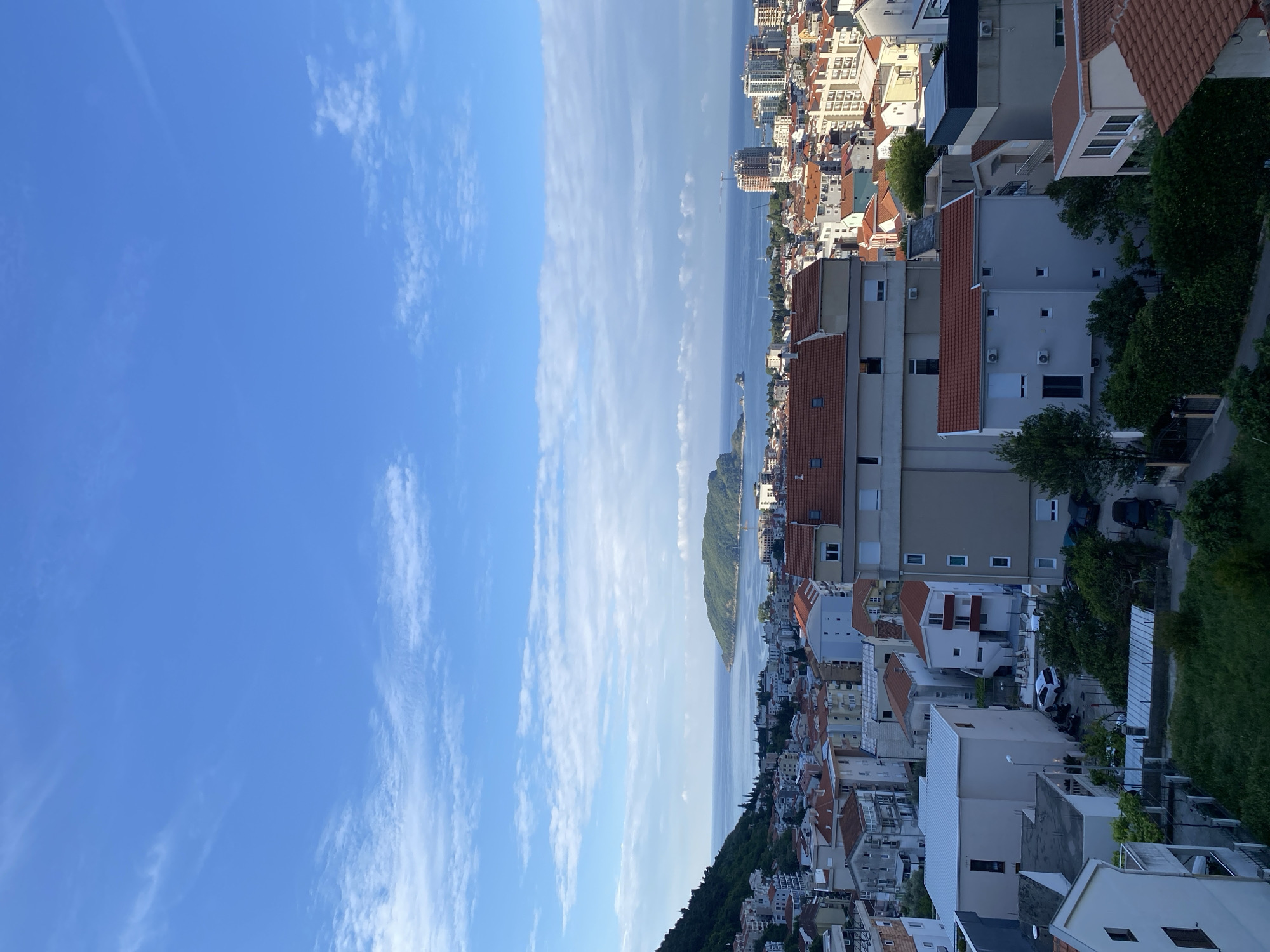 The image depicts a cityscape in Budva, Montenegro, likely photographed from an elevated position. The sky takes up a significant portion of the left side, with scattered cirrus clouds and clear blue sky. The right side shows densely packed buildings, with red tile roofs and light-colored walls, nestled along the coastline. In the distance, a mountainous island is visible in the water. The sea stretches horizontally between the sky and the city, with a few boats visible. The coastline features both urban development and green vegetation.
