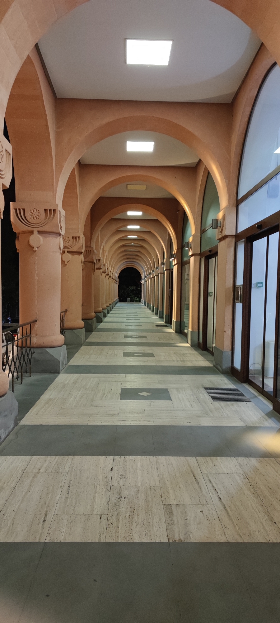 The image shows a long, covered walkway with a series of arches supported by substantial columns. The walkway is paved with rectangular light-colored stones interspersed with darker grey bands, creating a striped pattern that recedes into the distance. Several square or rectangular lights are embedded in the ceiling of the walkway, illuminating the scene. On the right side of the walkway are glass doors and windows, suggesting the walkway is connected to indoor spaces. On the left, there are railings and a dark background, possibly indicating an outdoor area or building exterior. The architecture features ornate carvings on the columns. The overall impression is one of a formal and well-maintained public space. This walkway is located at the Cascade complex in Yerevan, Armenia. There are no people visible in the image.
