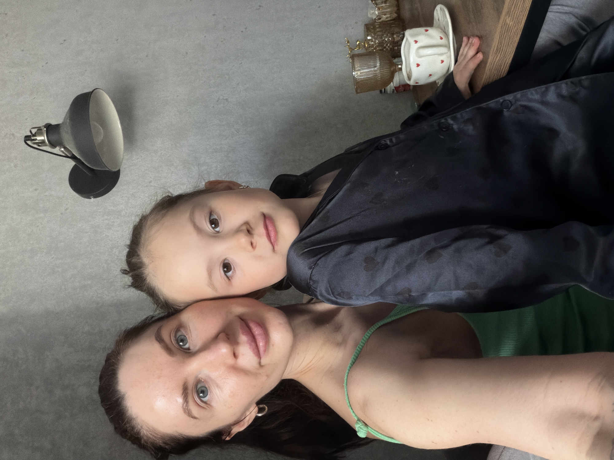 A selfie photograph features a woman and a young girl, possibly in Perm, Russia. The woman, with dark hair and light eyes, is wearing a green garment. Above her is the girl, with lighter hair and brown eyes, wearing a dark blue satin shirt. A lamp is visible in the upper left, and a wooden surface with glassware and a cup is in the upper right.