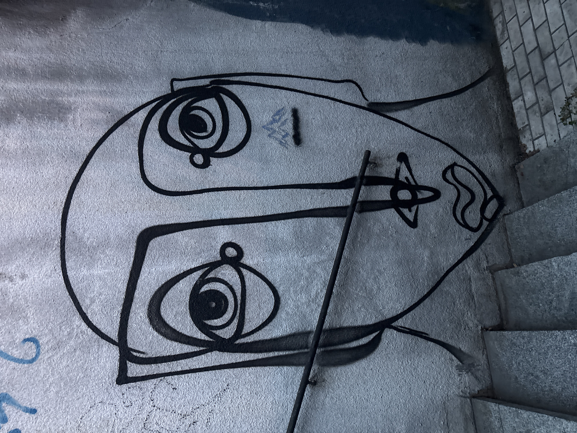 The image shows a section of a concrete wall with graffiti. The graffiti is in black outline and depicts a stylized face with two large eyes. The eyes are concentric circles with smaller circles inside, reminiscent of owls or abstract art. The face has a long, somewhat elongated oval shape. A thin, vertical black pole or stick runs across the middle of the face. To the right of the graffiti, a set of stone stairs is visible, suggesting an outdoor or semi-outdoor location. The lighting appears to be dim, possibly indicating early morning, late evening, or an overcast day. The concrete wall has a rough texture, and there are some lighter gray and dark blue smudges above the graffiti, possibly from other graffiti or environmental elements. There are also faint blue markings on the wall to the left of the main graffiti, and some faint scribbles below the face. The overall impression is that of urban street art.