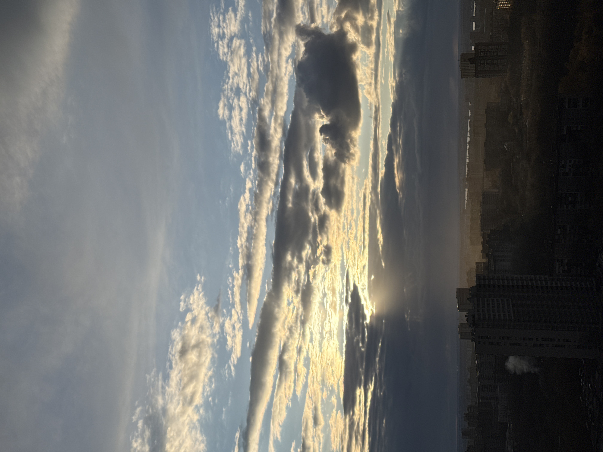 A panoramic landscape photograph captures a dramatic sky over a city, indicating either sunrise or sunset. The upper two-thirds of the image are dominated by an expansive sky filled with various cloud formations. Long, streaky clouds, likely stratocumulus or altocumulus, are brightly illuminated by golden sunlight, appearing in vibrant shades of white, yellow, and orange, contrasting with areas of light blue sky. Towards the right side, darker, denser clouds partially obscure the sun, from which striking crepuscular rays (sunbeams) pierce through, fanning out across the horizon.
The lower third of the image features a city skyline, largely silhouetted against the radiant sky. Tall, dark buildings are discernible, particularly on the right side of the frame, suggesting an urban environment. The distant landscape appears hazy and extends broadly beneath the sky, hinting at a vast urban or semi-urban area. The warm, intense lighting and deep shadows cast by the clouds and buildings are characteristic of either dawn or dusk. No visible text is present in the image. The location is Perm, Russia.