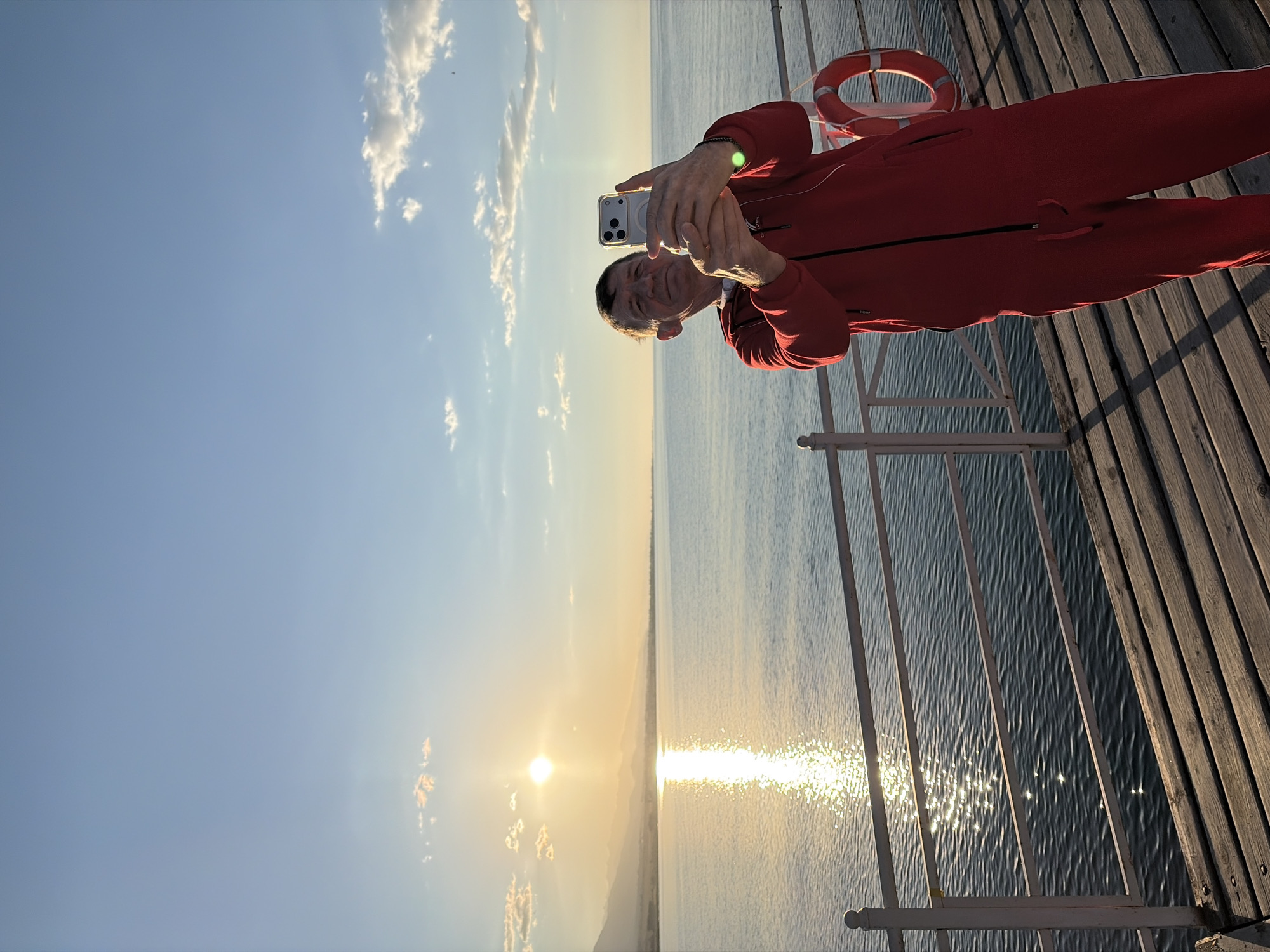 An adult male is standing on a wooden pier, taking a selfie with his phone. He is wearing a red tracksuit. The sun is setting over a large body of water, casting a golden reflection on the surface. Mountains are visible in the distance on the horizon. A life preserver is attached to the railing of the pier. The sky is blue with a few scattered clouds.