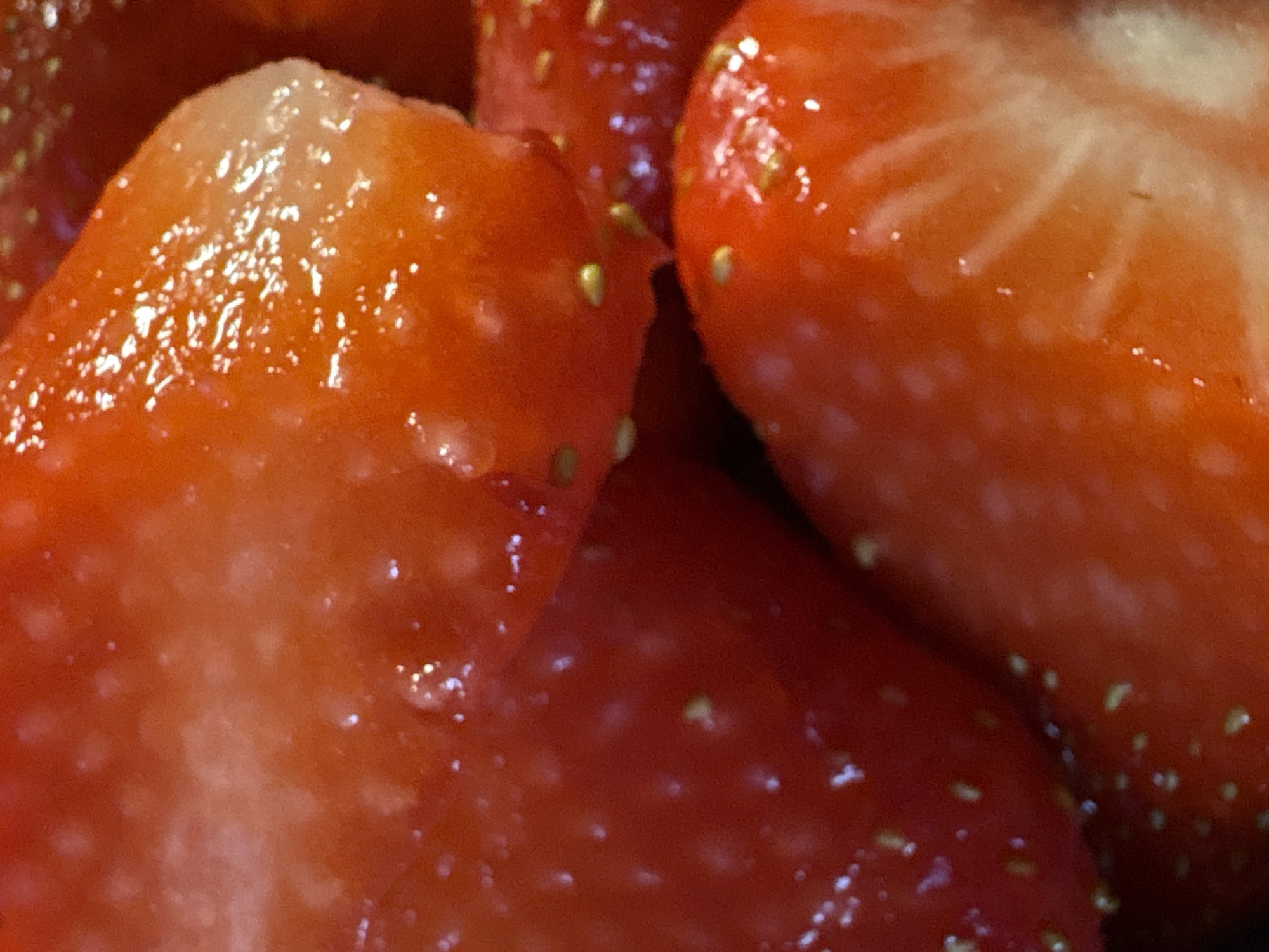 This close-up, macro photograph focuses on several vibrant red strawberries, filling the entire frame with their intricate details. The strawberries appear to be either whole or partially cut, revealing their contrasting lighter pinkish-white flesh beneath the glossy, deep red exterior. Numerous small, pale yellow or greenish seeds are visible embedded across the surface of the fruit. The strawberries glisten with moisture, indicated by many bright, specular highlights scattered across their textured surfaces, suggesting freshness and juiciness. The composition is tight, eliminating any discernible background or external setting, making the image almost abstract and emphasizing the fruit's natural beauty and texture. There are no people, explicit activities, or visible text within the frame. While no location cues are present in the image itself, the context provided specifies the location as Saint Petersburg, Russia. No particular time of day, weather conditions, or emotions are conveyed.