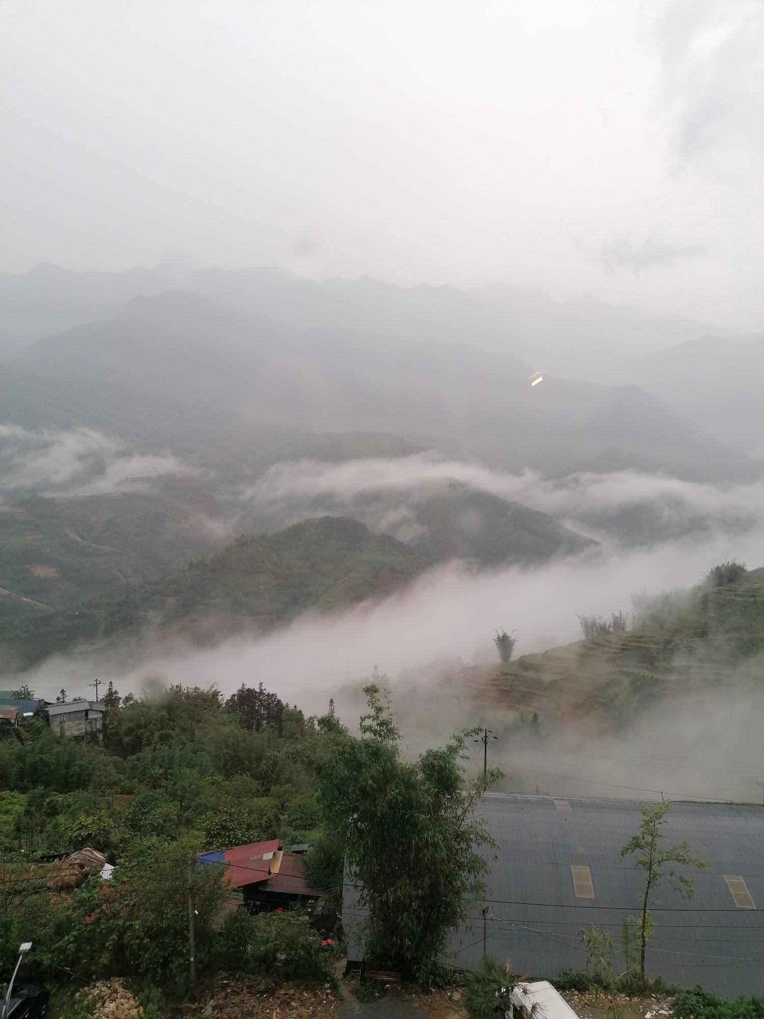 The image depicts a mountainous landscape in Sa Pa Ward, Vietnam, shrouded in heavy fog and mist. The mountains are visible in the background, their peaks partially obscured by the low-lying clouds. In the foreground, there is vegetation, including trees and shrubs, and some buildings with red and grey roofs. There's also a large grey building with skylights and some power lines. The overall atmosphere is overcast and damp.