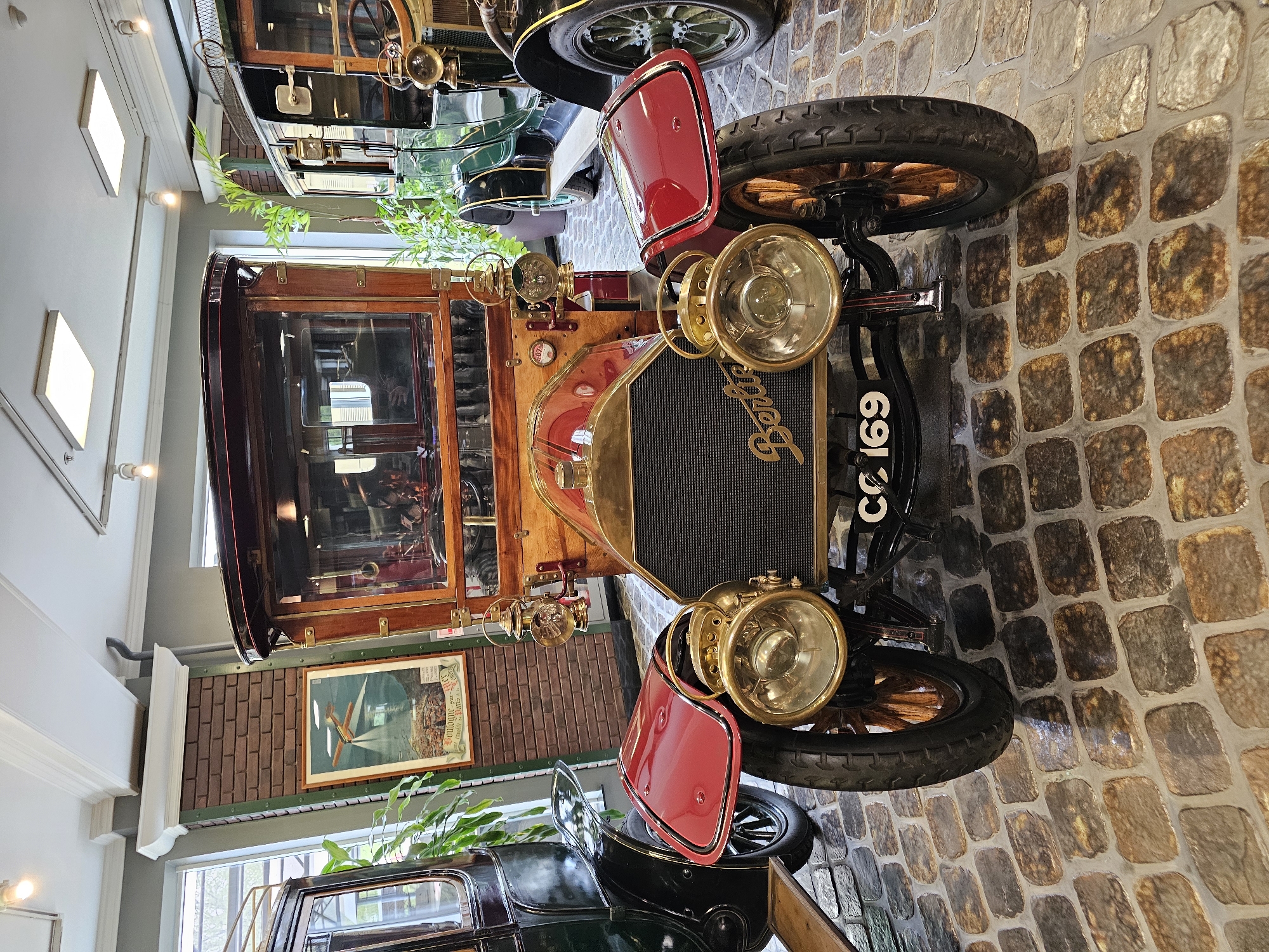 A vintage "Berliet" automobile is displayed inside a building. The car features a wood-paneled body, red fenders, brass headlamps, and a black grille with the Berliet logo. Its license plate reads "CC 169". The car is positioned on a cobblestone floor. In the background, there's another classic car, a framed picture on a brick wall, plants, and architectural details of the building's interior. The image was taken indoors, likely in the Архангельское, Russia car museum.