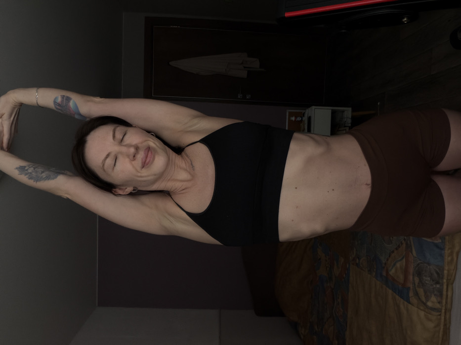 The scene depicts an indoor residential setting in Perm, Russia. A single adult female stands centrally, facing forward, with her eyes closed and a relaxed expression. She performs a full-body stretch, extending both arms overhead with hands interlocked. She is attired in a black sports bra and dark brown athletic shorts. Visible tattoos include a stylized feather or bird design on her upper left arm and a colorful, multi-toned feather or abstract design on her upper right arm. She wears small silver hoop earrings in both ears and a thin silver bracelet on her right wrist. Her abdomen shows distinct rib definition.
The background to the left reveals a bed with a dark headboard and a patterned duvet in shades of brown, beige, and blue. To the right, a dark wooden door with a silver-colored handle has a light-colored garment, possibly a robe or towel, hanging from a hook. Adjacent to the bed, a white nightstand is partially visible, holding a small white bottle with an orange cap. On the far right edge, a dark object with a prominent red stripe, consistent with exercise equipment, is partially visible. The room's walls are a dark, muted color, possibly dark purple or gray. The lighting is dim and soft, suggesting a private space such as a bedroom.