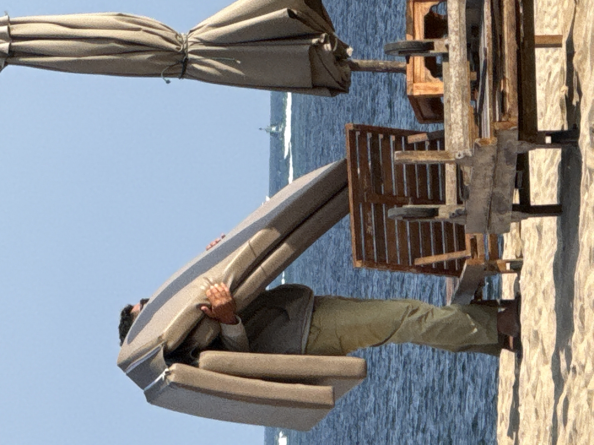 A man stands on a sandy beach, holding a folded beach chair. He is wearing a light-colored long-sleeved shirt and khaki pants. The beach chair is made of a light-colored fabric and has a dark stripe. In the background, the ocean is visible, with a small boat in the distance. A large umbrella is deployed to the right of the frame, and another beach chair is partially visible in the foreground. The scene appears to be in Denpasar, Indonesia, possibly on Kuta Beach.
