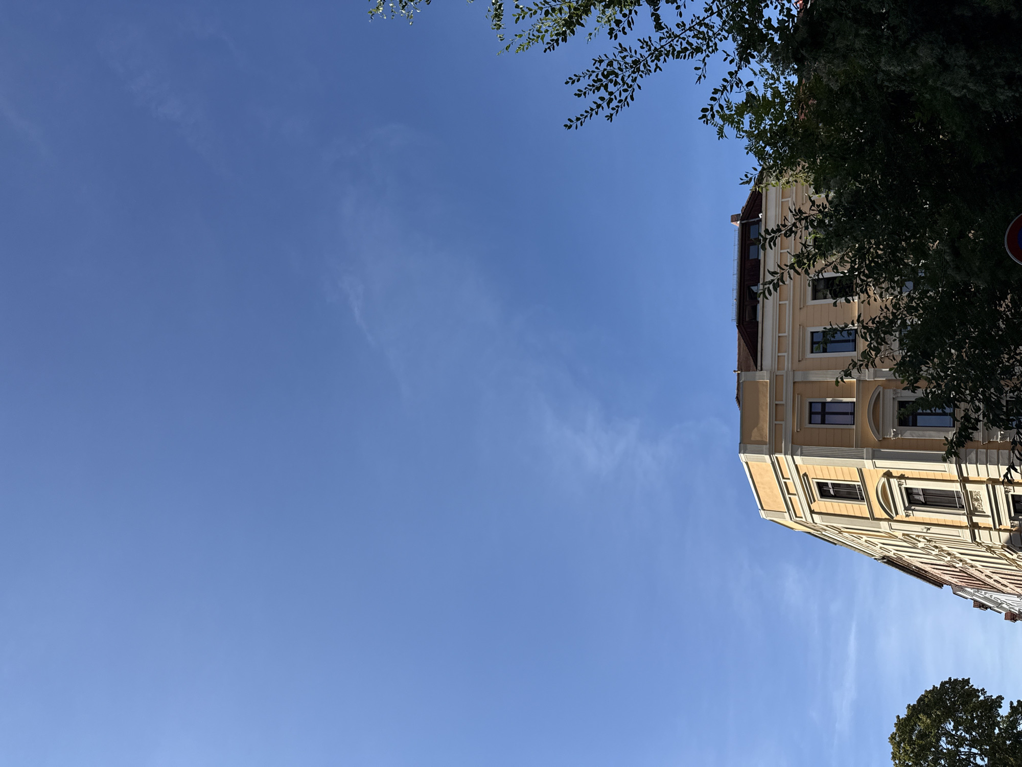 A bright blue sky with thin wisps of clouds dominates the upper portion of the image. In the lower half, a multi-story yellow building with an ornate facade, characteristic of the Gründerzeit architecture found in Görlitz, Germany, is visible. The building has numerous windows with dark frames and features classical architectural elements like columns and cornices. To the left and right of the building, dense green foliage from trees partially obscures the view. The sunlight suggests it is daytime.