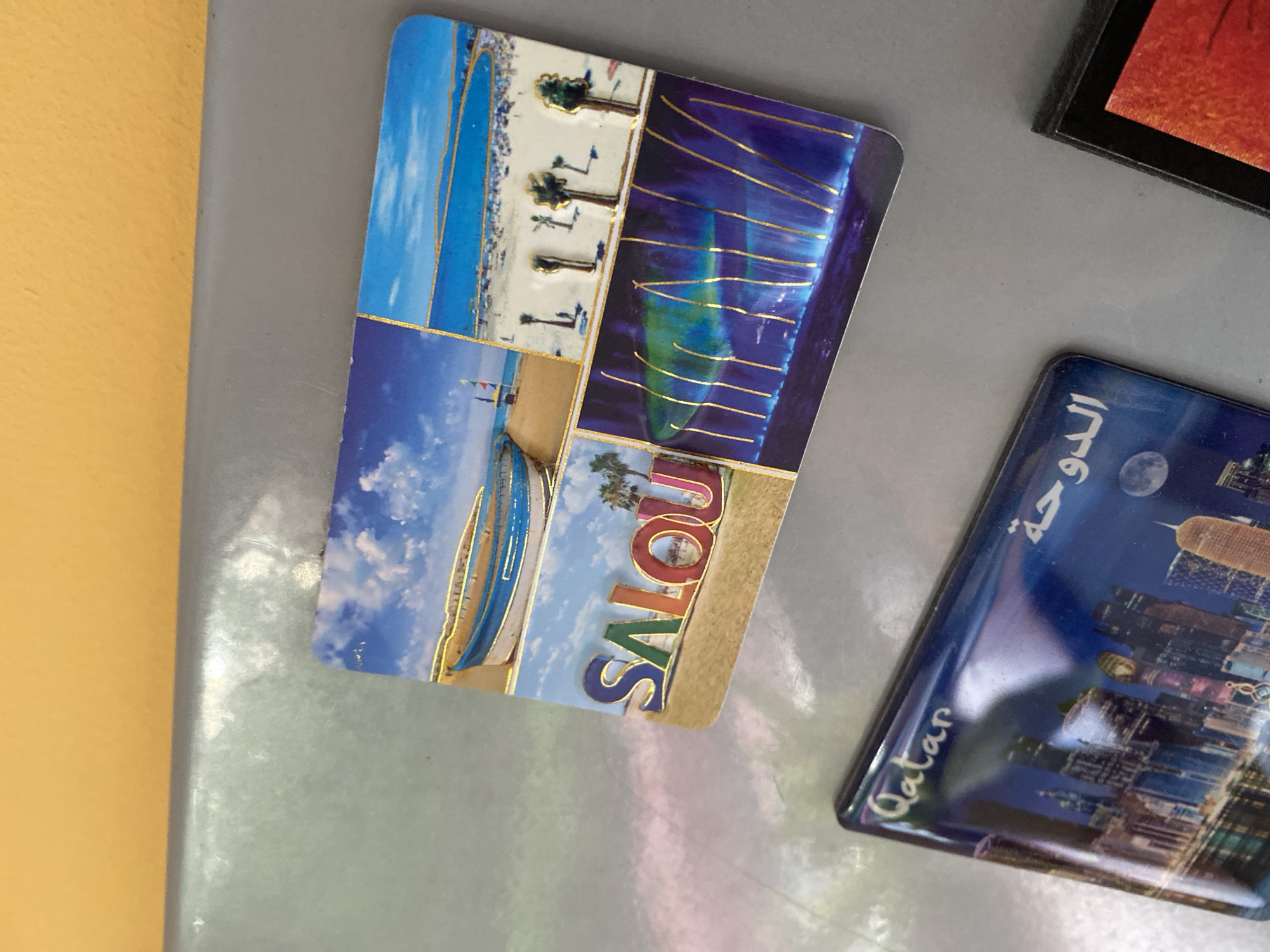 The image shows a close-up view of several refrigerator magnets attached to a metallic surface, likely a refrigerator door. The magnets are colorful and feature various scenes.
The top magnet is a collage of four images. The top left shows two traditional boats pulled onto a sandy beach under a blue sky with clouds. The top right depicts a beach scene with people, palm trees, and buildings in the distance, also under a blue sky. The bottom left features the word "SALOU" in large, colorful, three-dimensional letters, with a tropical scene including palm trees and a lighthouse in the background. The bottom right shows an abstract depiction of colorful lights or a fountain at night, with golden lines overlaying the image.
Below the collage magnet is another magnet with a nighttime cityscape of tall buildings. The word "Qatar" is written in script on the left side, and Arabic script appears on the right.
To the right of these magnets, a portion of another magnet or item is visible, displaying a vibrant orange and red abstract pattern.
The metallic surface appears to be light gray with some subtle reflective qualities, showing slight discoloration or wear. The background above the metallic surface is a solid, bright orange color.
There are no people visible in the image, and no specific objects or details suggest the location is Wrocław, Poland. The magnets depict travel destinations.
