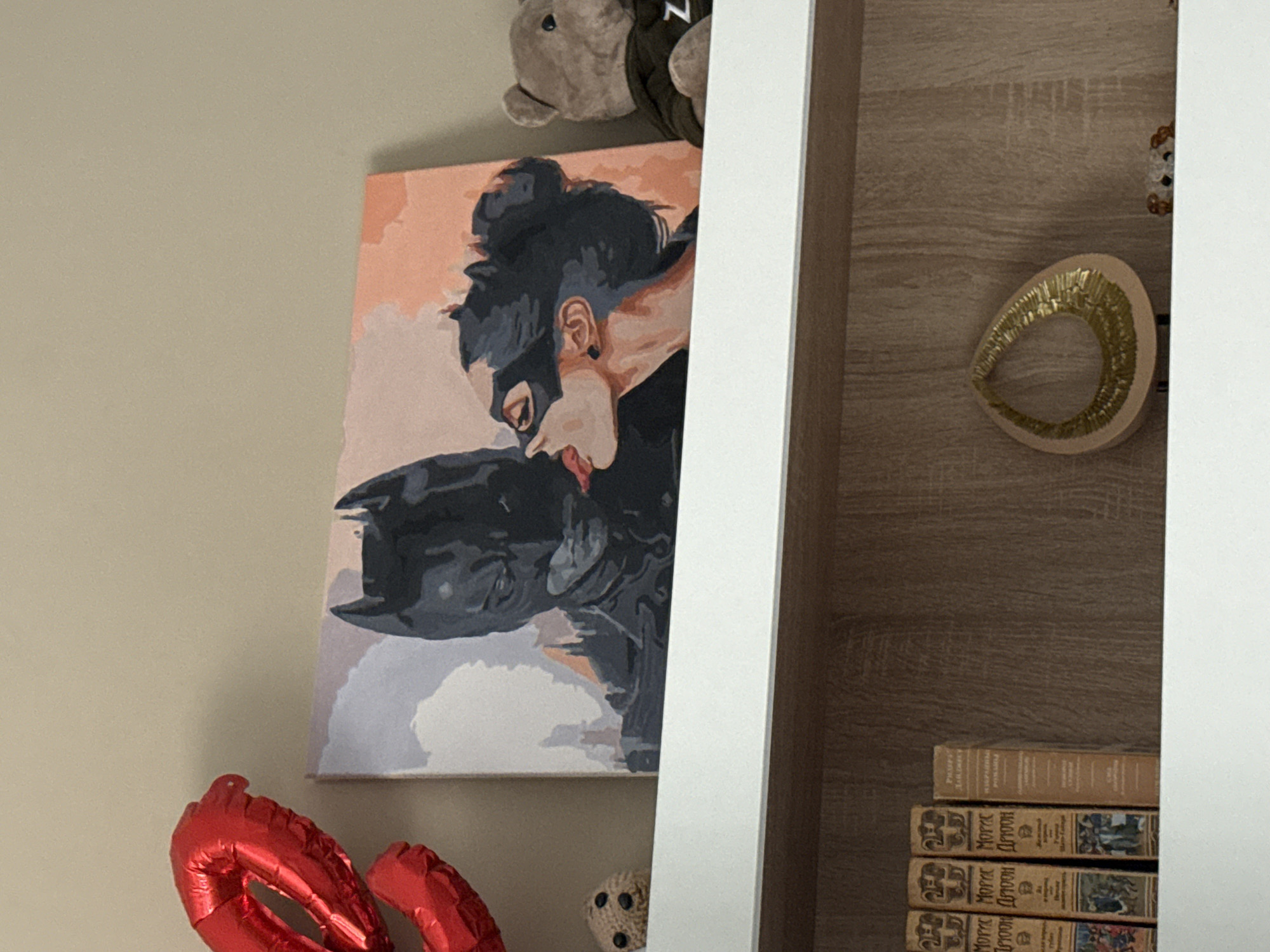 The image shows a painting of Batman and Catwoman kissing, hung on a wall. Next to the painting is a wooden shelf unit. On the shelf are a golden egg-shaped ornament, a stuffed animal, and a stack of books. Below the painting are a red balloon and a small stuffed ball. The scene is located in an unknown city.