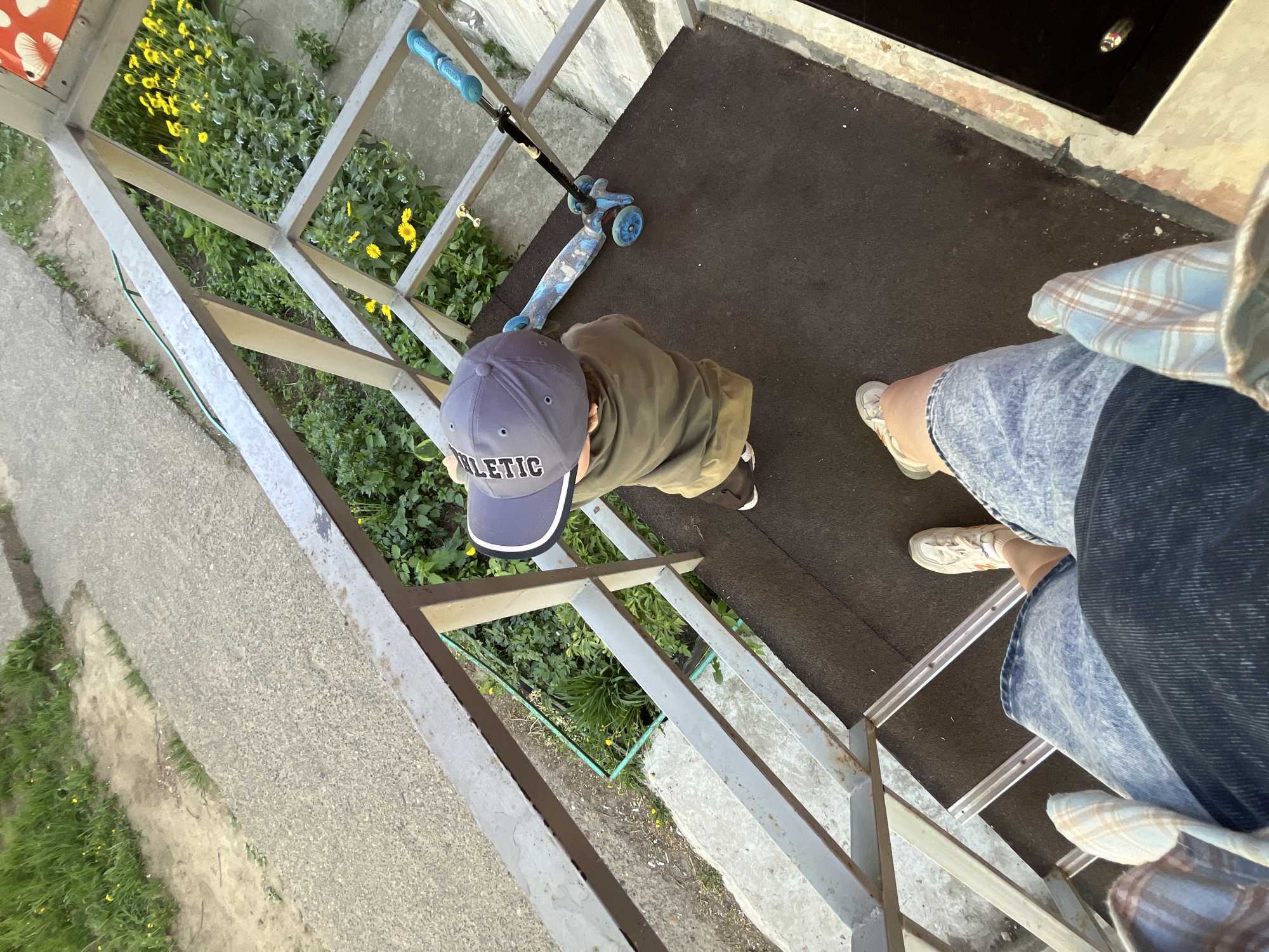 A high-angle shot shows a child and an adult on a walkway in Chaikovsky, Russia. The child, wearing a grey cap with the word "ATHLETIC" on it, a green jacket, and black shoes, stands near a blue scooter on a black rubber surface. The adult, wearing blue jeans, a plaid shirt, and white sneakers, stands near the edge of the walkway. The walkway is made of metal and overlooks a grassy area with yellow flowers and a concrete path.