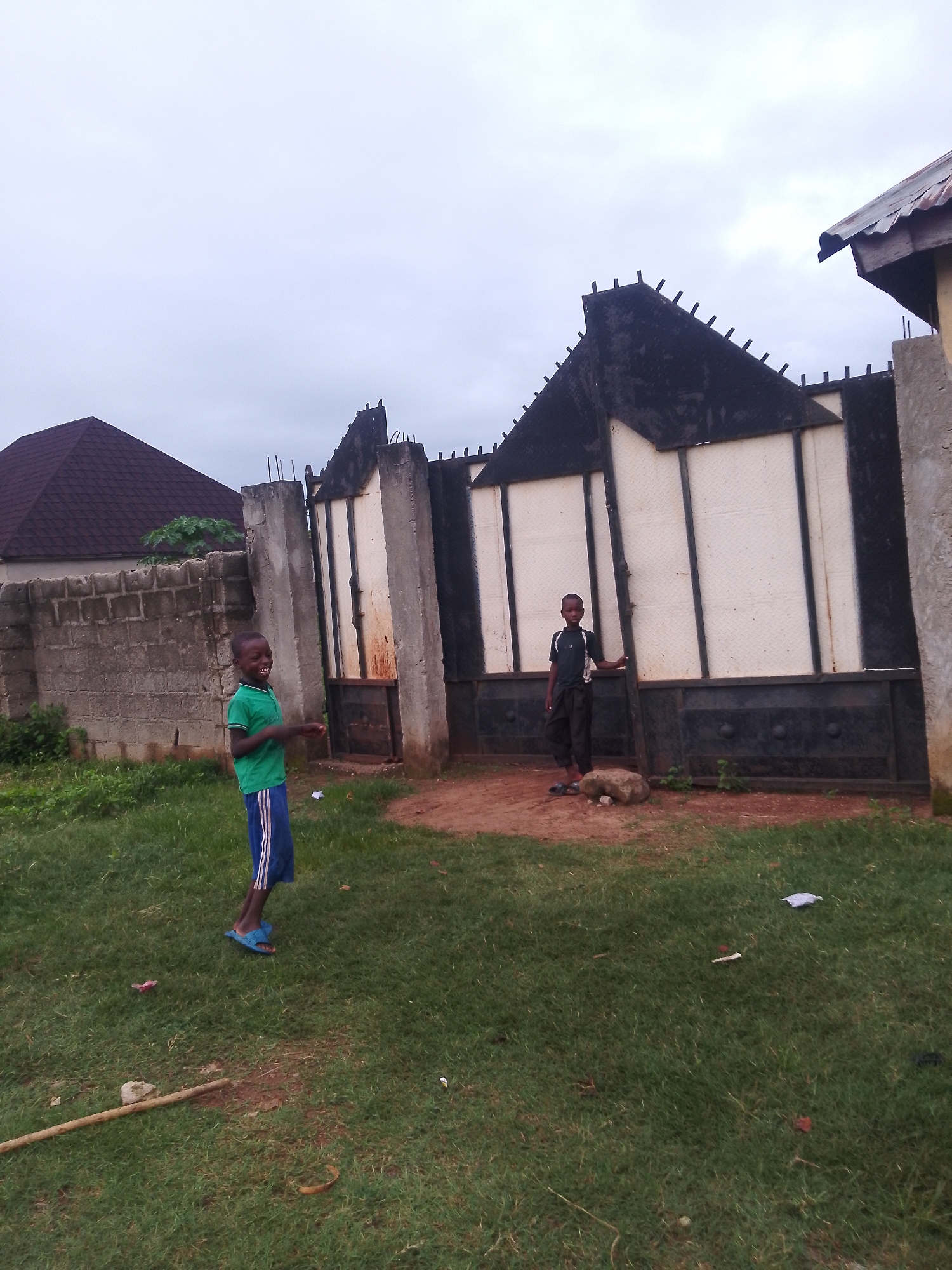 In Serti, Nigeria, a boy in a green shirt and blue shorts with white stripes, wearing blue sandals, stands on a grassy area, facing left and smiling broadly. He appears to be looking at something off-camera. Behind him, to the right, another boy in a dark shirt and dark pants stands leaning against a large, decorative metal gate. The gate is segmented with white panels and dark framing. To the left of the first boy is a section of a concrete block wall. Beyond the wall, a house with a dark, patterned roof is visible. The environment consists of grass, dirt, and a cloudy sky.