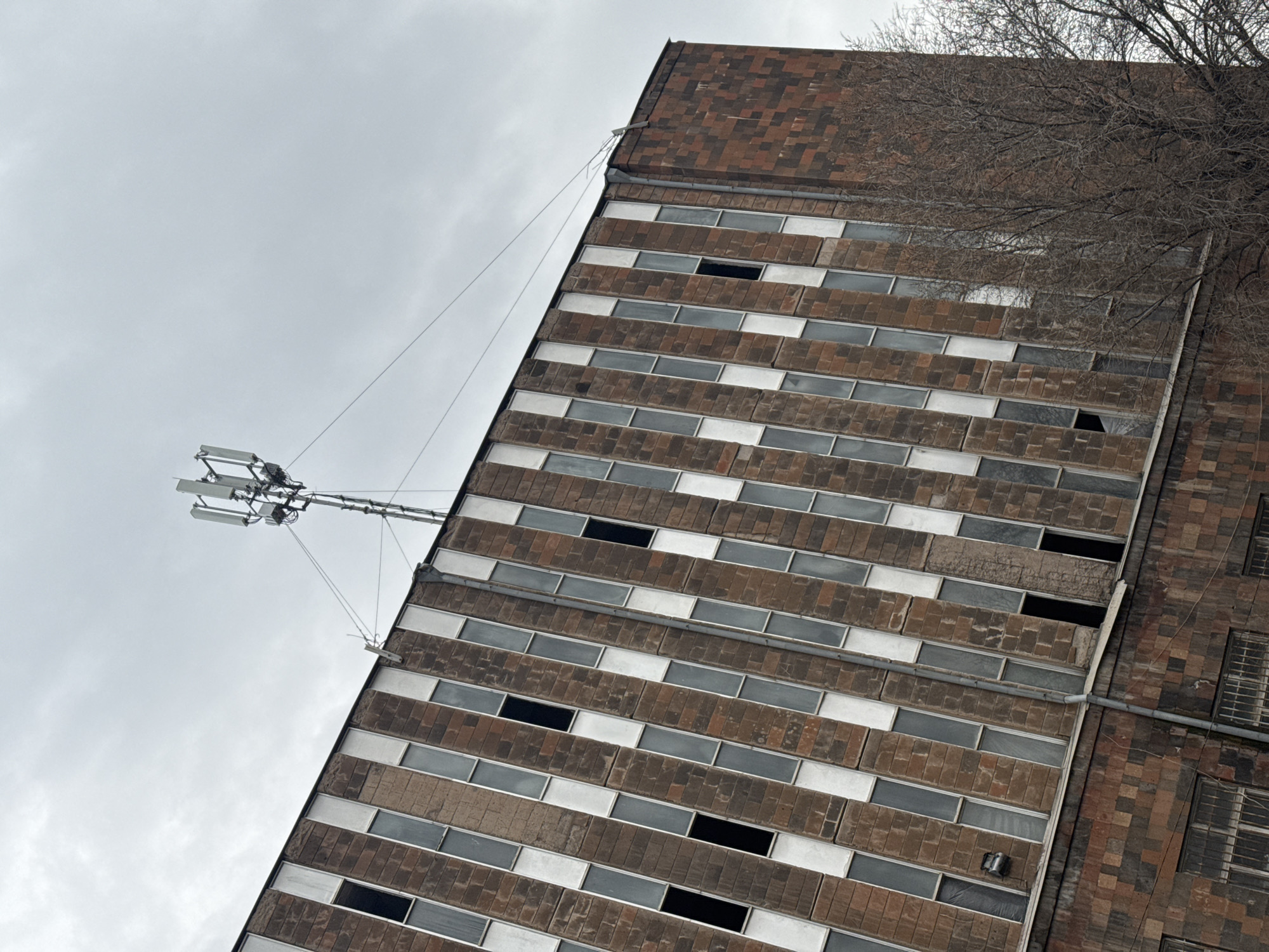 The image depicts the exterior of a multi-story building, possibly in Yerevan, Armenia, though this is unconfirmed. The building is constructed from brown brick and concrete with a repetitive pattern of long, rectangular windows. Some windows appear to be missing. A communications tower or antenna is attached to the building's edge via cables. The sky is overcast and grey. A bare tree is partially visible at the top right corner of the image.