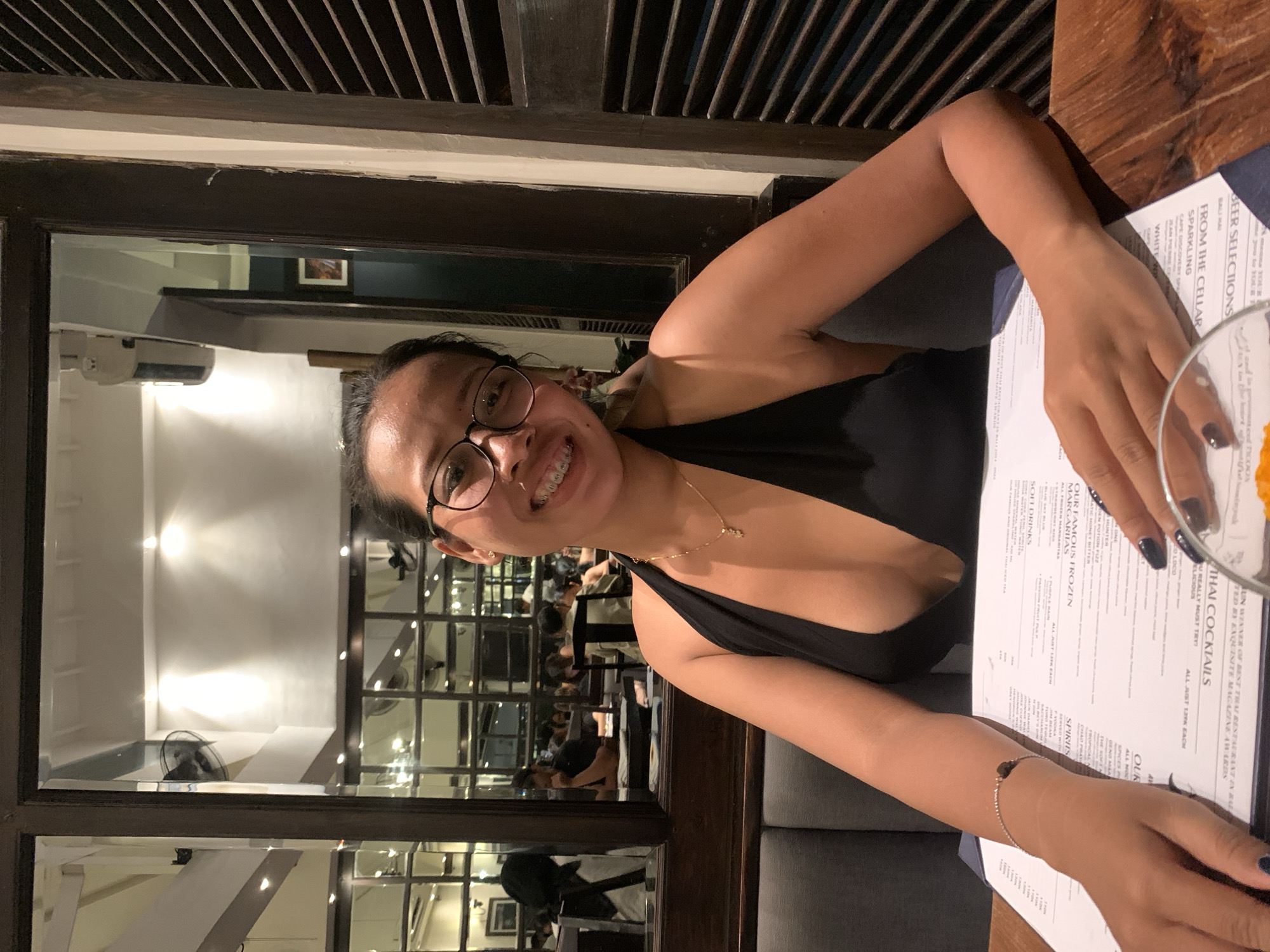 A person with glasses and braces smiles at the camera while seated at a table in what appears to be a restaurant. The restaurant is located in Kuta, Indonesia, according to a menu on the table. The person is wearing a black top and has a gold necklace and bracelet. A cocktail and the menu are on the table in front of them. A mirror behind them reflects the interior of the restaurant, showing other patrons and staff.