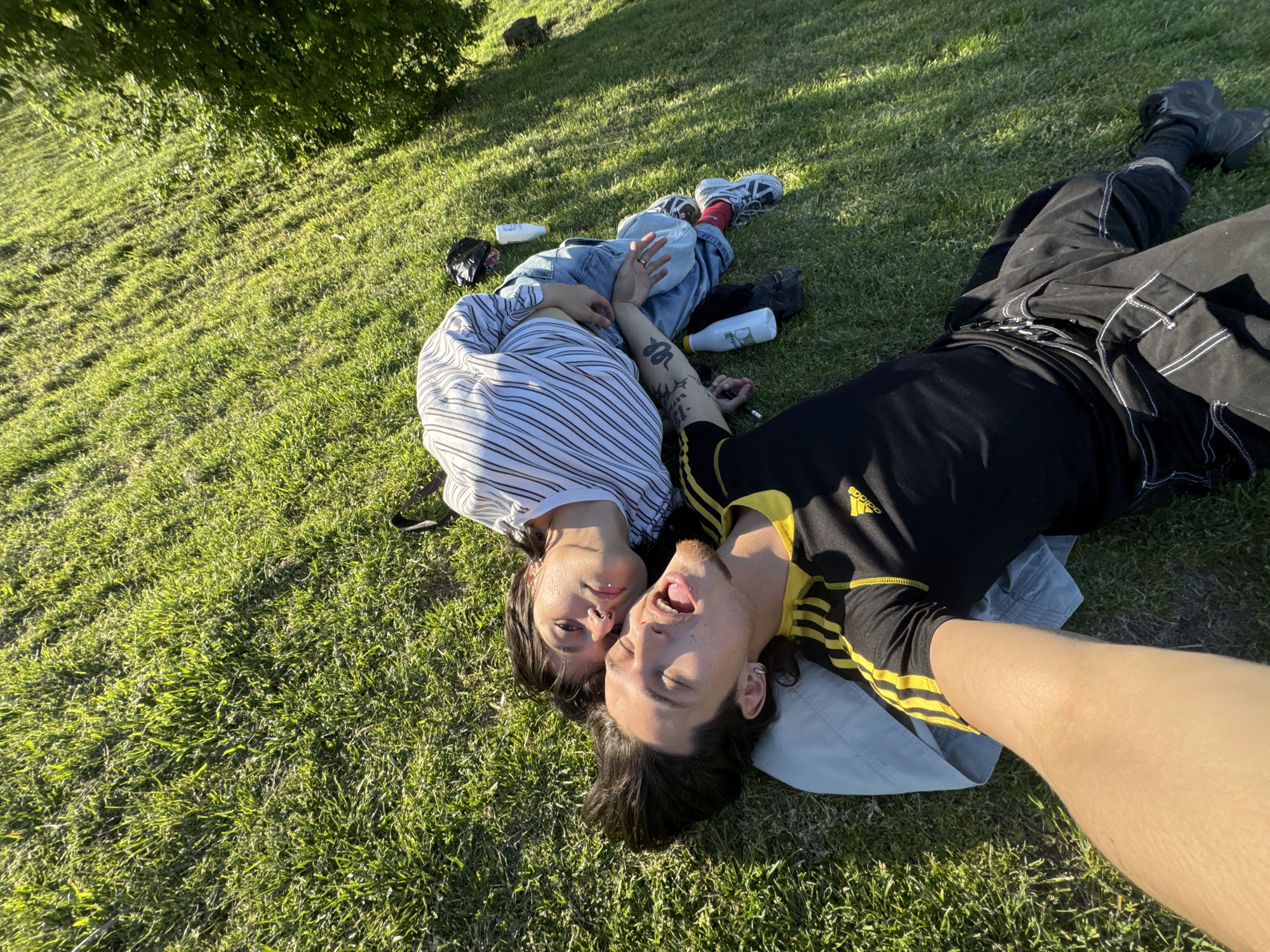 Two people are lying on a grassy surface. The person on the left wears a striped shirt and jeans and is positioned with their head close to the other person's face. The person on the right wears a black Adidas shirt with yellow stripes and black pants with white stitching. They are lying on a pillow. There are water bottles and shoes scattered around them. The location appears to be a park, based on the grass and trees visible in the background. Sofia, Bulgaria cannot be determined based on the visual information.