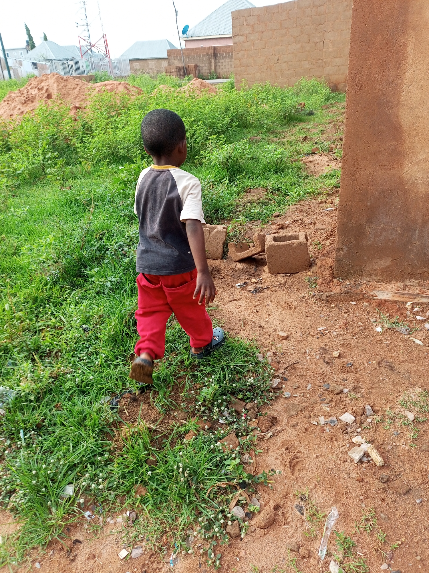 In a residential area of Tempure, Nigeria, a young boy wearing a dark t-shirt and red pants walks away from the camera on a dirt path. The path is bordered by green grass. In the background, several houses are visible, along with piles of sand and a red metal tower. The boy is wearing dark sandals. The scene appears to be a sunny day.
