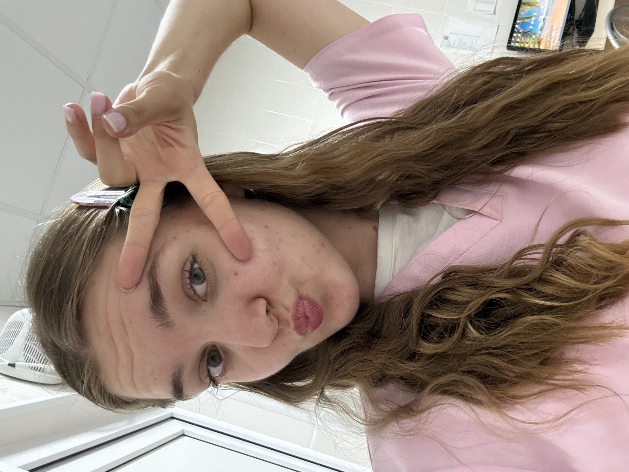 A young woman with long, wavy brown hair is taking a selfie. She is wearing a pink scrub top over a white shirt. Her hair is partially held back by a green and pink hair clip. She is puckering her lips and holding up two fingers in a peace sign. Her eyes are green and she has light skin with some freckles. The background appears to be an indoor room with white walls and a window to the left. A computer monitor is visible in the background on the right. The lighting suggests an indoor setting during the day. There are no specific landmarks or indicators of the city of Perm, Russia, in the image.