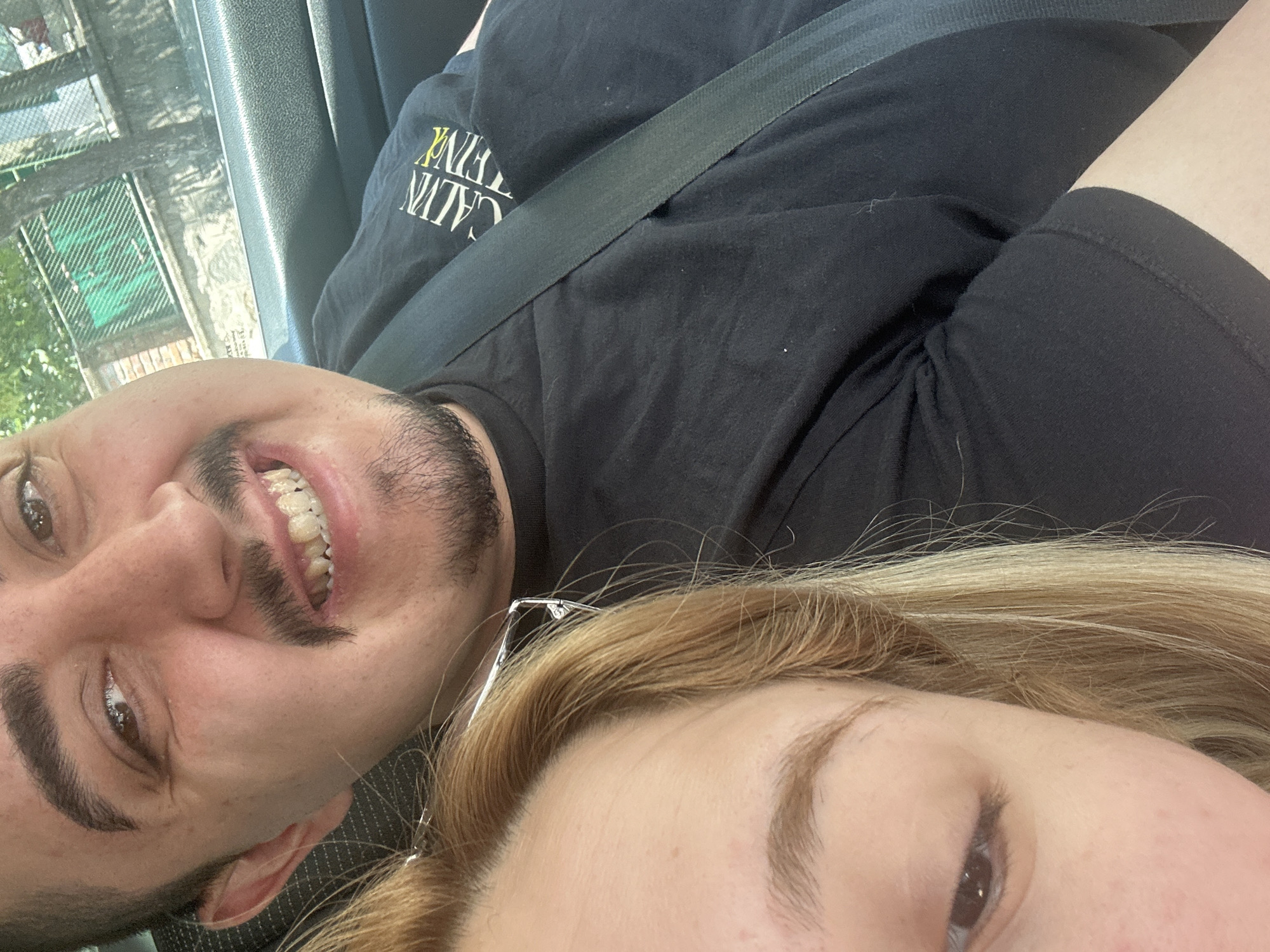 The image is a close-up, selfie-style photograph taken from inside a vehicle, likely a car. In the foreground, a person with blonde hair and fair skin is partially visible on the left, their eye and forehead in focus. To the right, a person with brown eyes, a mustache, and a goatee is smiling, revealing visible teeth. They are wearing a black t-shirt with a logo that appears to say "CALVIN" in yellow and white text. A black seatbelt is prominently displayed across their chest. The background shows a blurred view of trees and a green fence, suggesting an outdoor setting, possibly in a residential or urban area. The lighting indicates it is daytime. Based on the overall appearance and context, it is not possible to definitively determine the exact location in the city of Plovdiv, Bulgaria from the image alone.