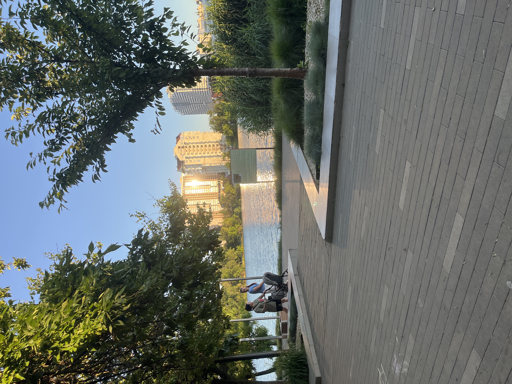 The image depicts a serene urban park setting with a body of water reflecting the warm glow of the setting sun. In the foreground, a paved walkway made of grey rectangular stones leads towards the viewer. To the left, a lush green tree with dense foliage partially obscures the view, while a lighter paved area with decorative stones and low-lying green plants can be seen.
In the middle ground, two individuals are visible on the walkway near the water's edge. One person, wearing a green top and dark shorts, is holding a bag. The other person, in a blue t-shirt and dark pants, is standing next to a stroller. They appear to be engaged in a casual stroll.
The background features a tranquil lake or river, with the sun's rays creating a shimmering effect on its surface. Across the water, a cluster of multi-story apartment buildings rises, bathed in the golden light of the sunset. A thin, vertical green signpost is also positioned near the water. The sky is a clear, pale blue, characteristic of a late afternoon or early evening. The overall scene suggests a peaceful recreational area within a city. This park is likely to be located in Krasnodar, Russia, given the architectural style of the buildings and the presence of trees common in the region.