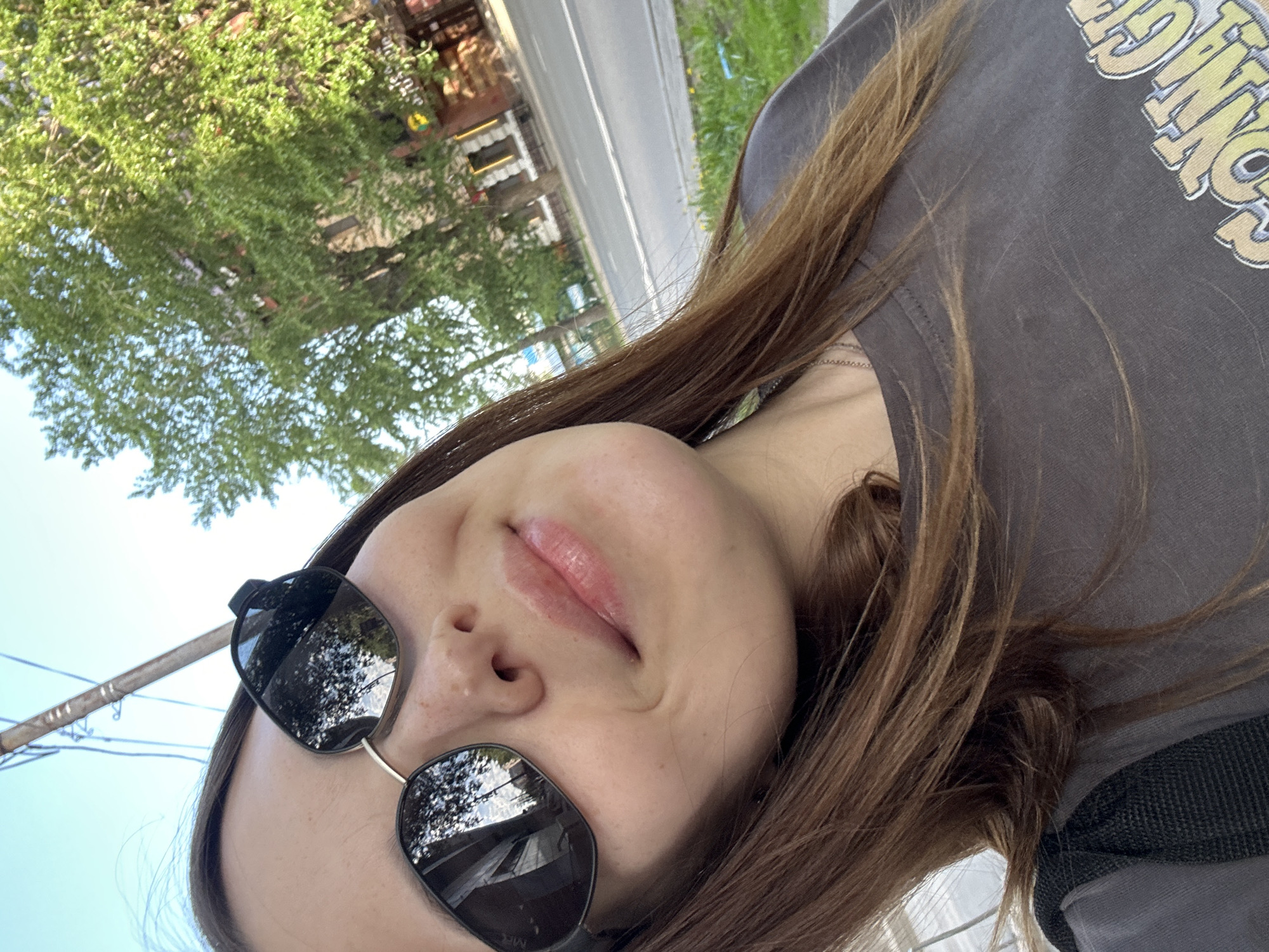 A selfie taken by a young woman in Perm, Russia, captures a scene with urban and natural elements. The woman is wearing sunglasses and a grey t-shirt. A tree with green leaves is visible in the background, along with a road and buildings. A utility pole is in the foreground near the top-left.