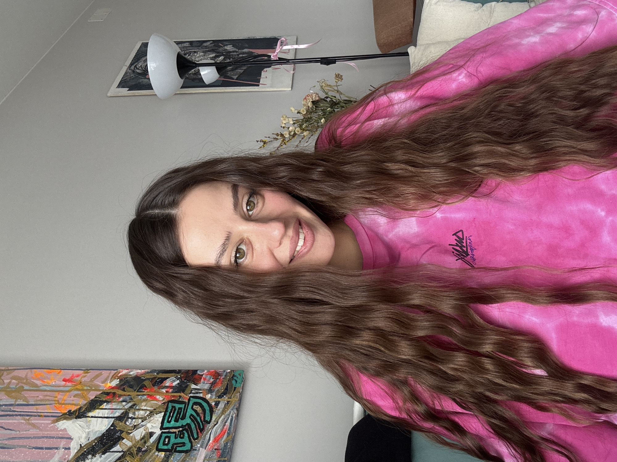 A medium shot captures a woman with long, wavy brown hair, smiling. She is wearing a pink tie-dye shirt. In the background, a gray wall is visible with a painting in the lower left corner. Further up, there's a lamp and another framed artwork. A bouquet of dried flowers is also present. A portion of a cushion is visible in the upper right corner. This image was taken in an interior setting, but the specific location in the city of Krasnoyarsk, Russia cannot be determined from the image.