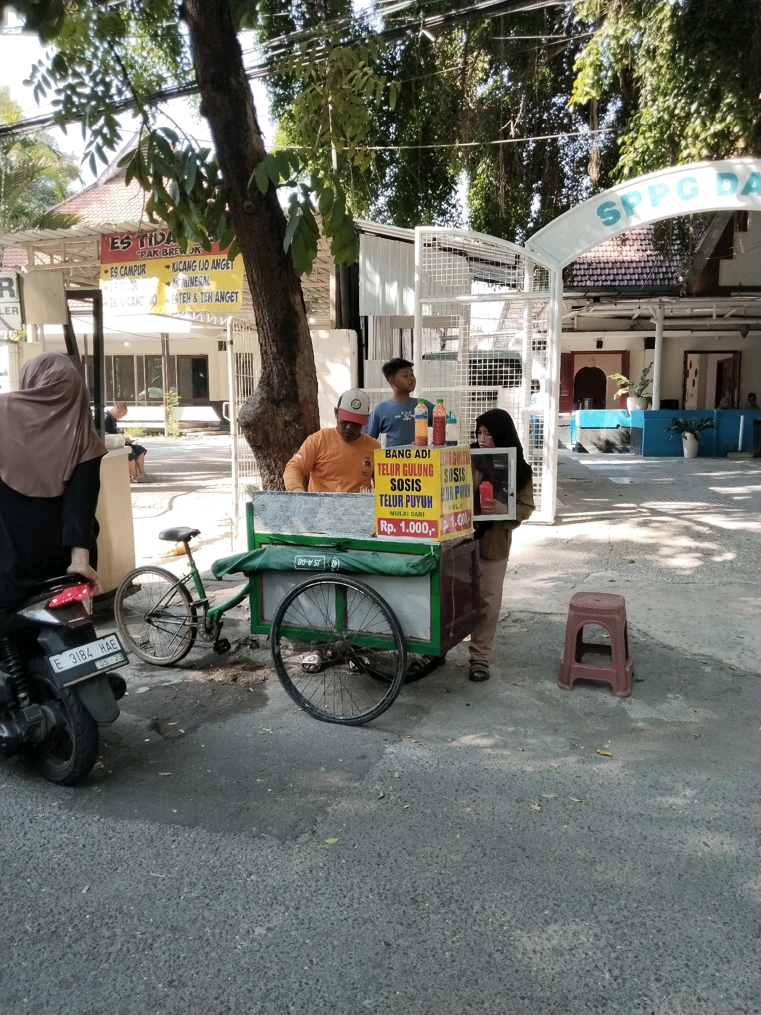 The image captures a street scene in Surabaya, Indonesia, likely in front of a building complex. In the foreground, a woman wearing a hijab stands next to a motorcycle. To her right, a food cart is situated, with a man in an orange shirt attending to it. A young boy in a blue shirt stands behind the cart. The cart displays various food items, including "TELUR GULUNG," "SOSIS," and "TELUR PUYUH," with prices listed. A bicycle is parked near the cart. The background shows a building with a tiled roof and a sign that partially reads "SPPG DA". Sunlight filters through the trees, casting shadows on the ground. The atmosphere appears to be that of a typical day in a bustling Indonesian city, with a focus on street food vending.