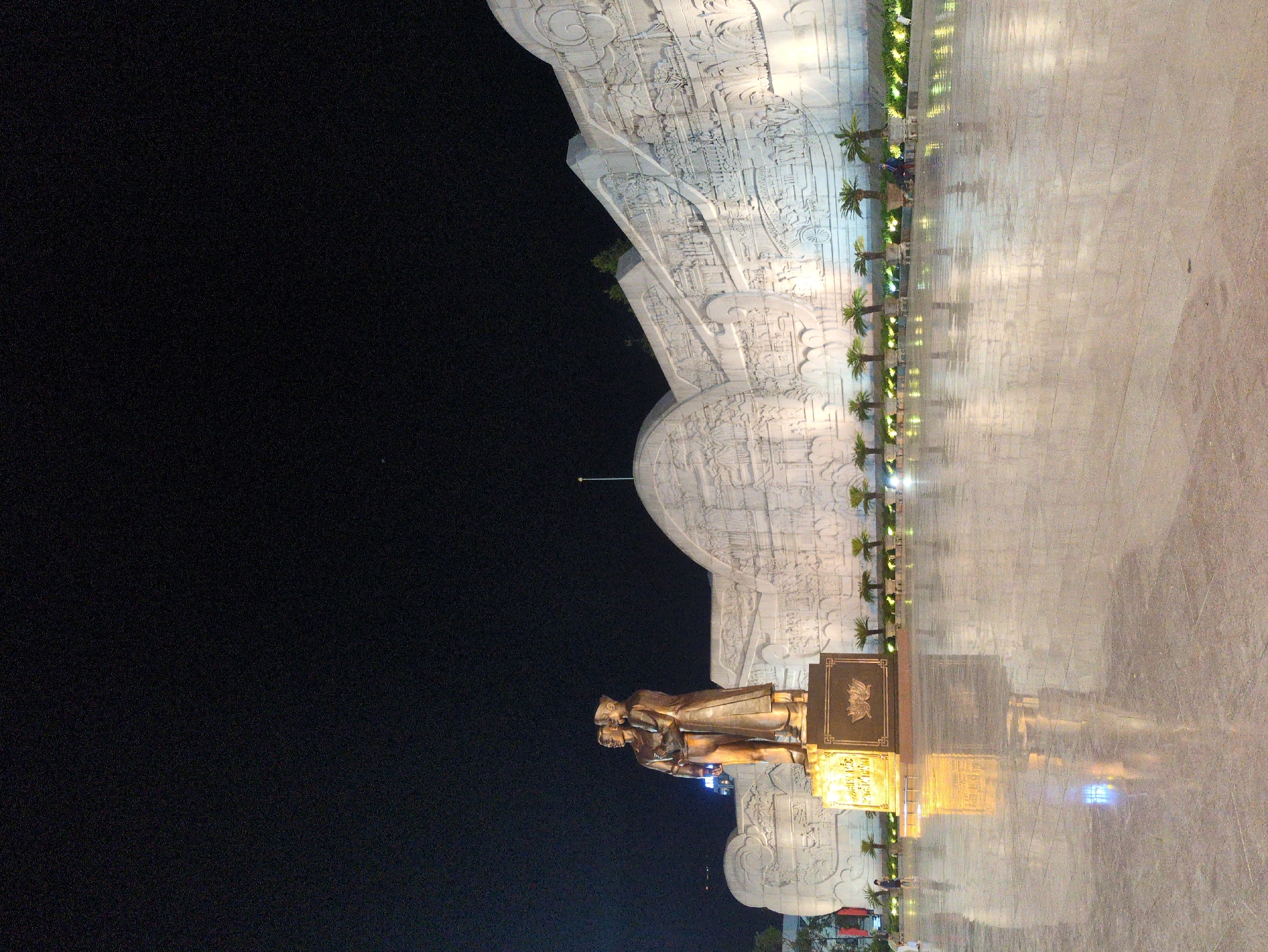 A vertical, nighttime shot shows a monument in Quy Nhon Ward, Vietnam. The monument consists of a bronze statue of two figures in Western attire, with a golden sign bearing Vietnamese text behind them. They stand in front of a large, elaborate, light-colored concrete relief. A row of small palm trees lines the base of the relief. The ground in front is a light, reflective surface. The sky above the monument is dark.