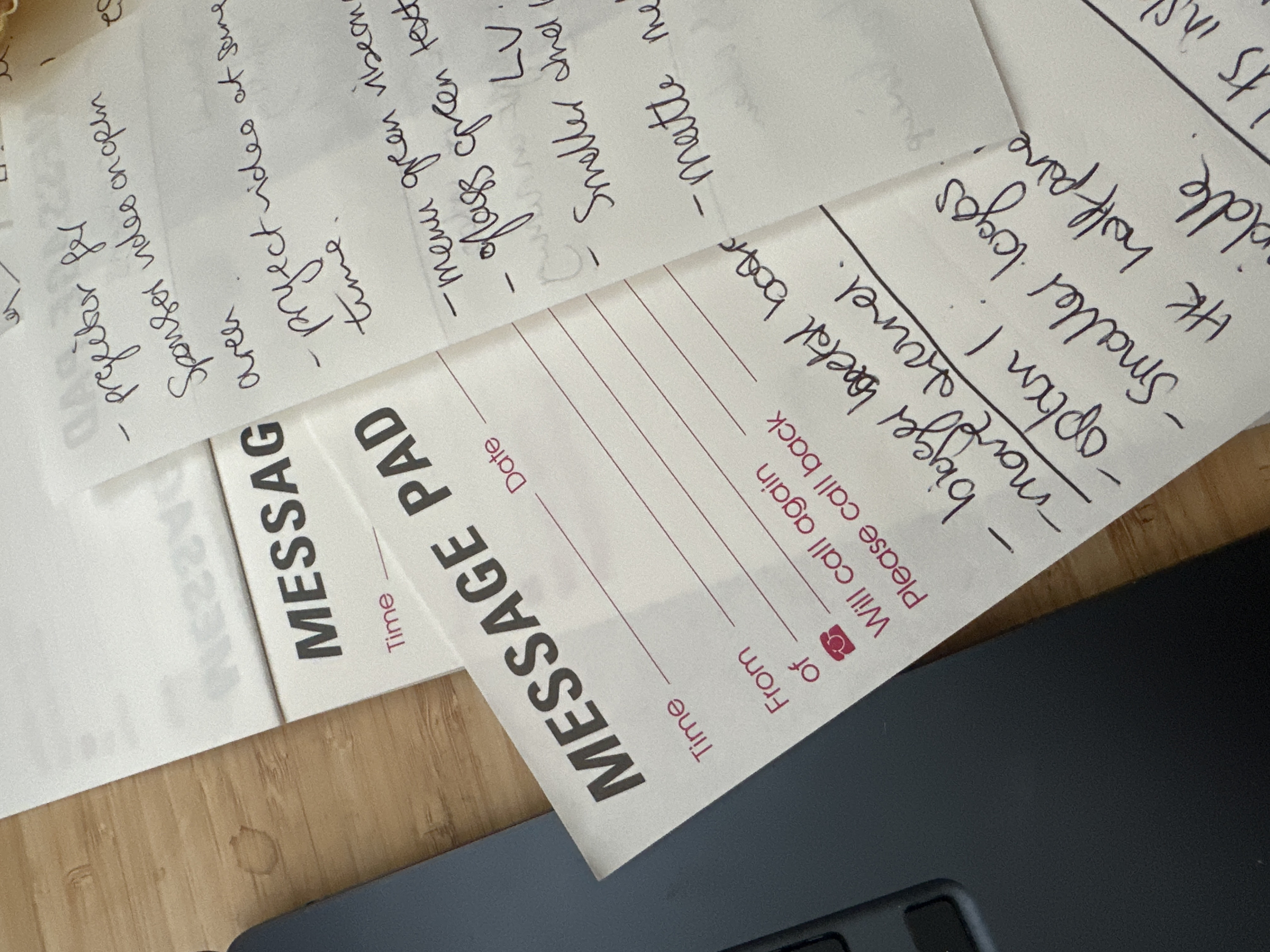 The image displays a close-up, overhead view of an indoor desktop surface in Tunbridge Wells, United Kingdom. Multiple white paper message pads, each bearing "MESSAGE PAD" in dark grey bold text and pre-printed fields like "Time," "Date," "From," "Of," and "Will call again" / "Please call back" in magenta ink, are casually stacked. Numerous handwritten notes in dark ink are visible across these papers. Examples of visible notes include: "- projector for", "Sponsor video on open", "area", "- project video at sime", "time", "- menu green ribbon", "- flags green text", "- Smaller and L", "- matte me", "bigger total boos", "- move kernel", "- option I", "- Smaller logos", "HK half pare", and "middle". A dark grey or black electronic device, consistent with the top-left corner of a laptop or a keyboard, is partially visible on the left side of the frame, resting on a light brown wooden surface. The wooden surface exhibits distinct grain patterns and several faint, circular liquid stains. No individuals or active interactions are present in the scene.