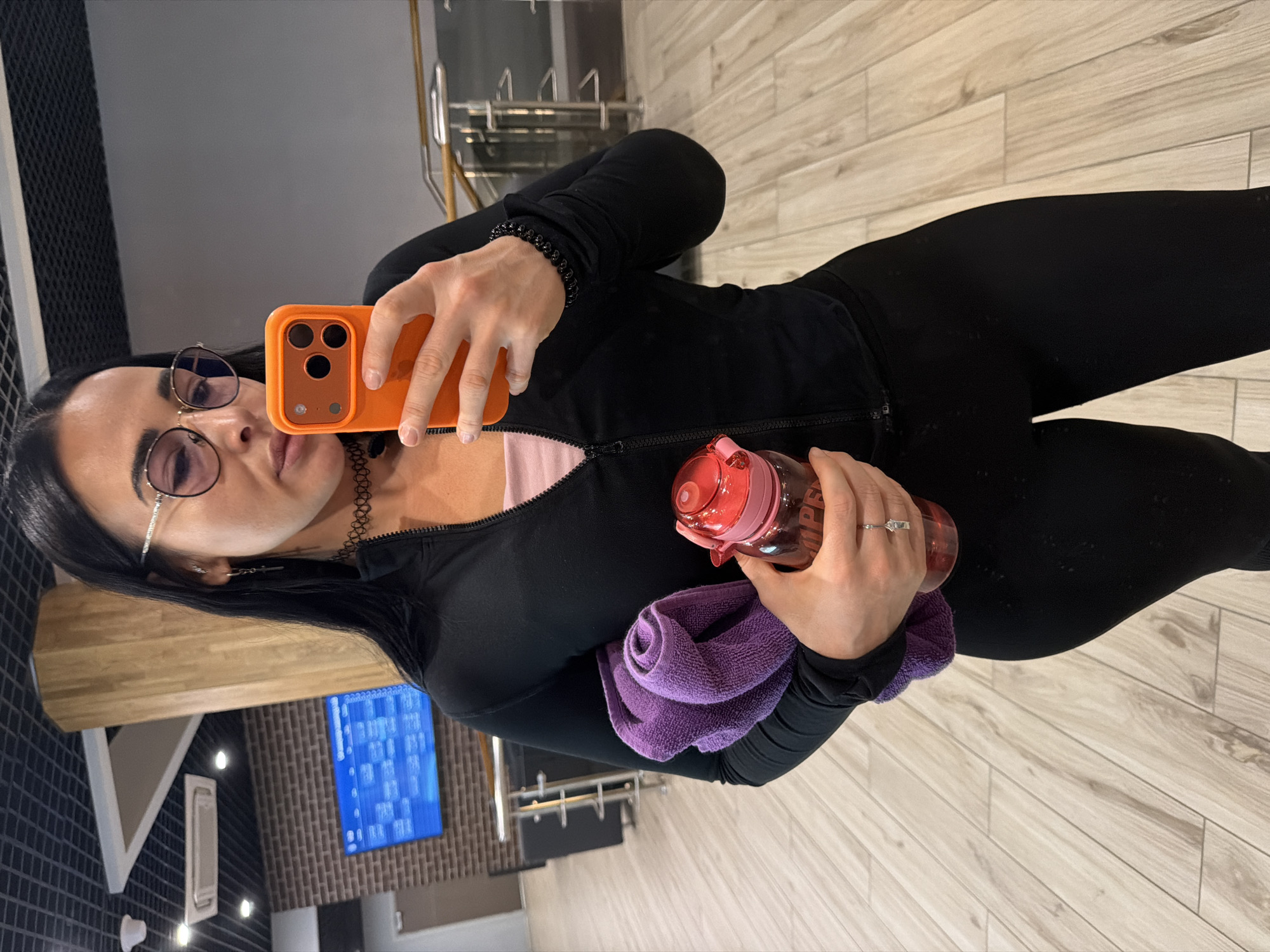 A woman is depicted taking a mirror selfie indoors, likely within a gym or fitness center. She has dark hair and is wearing round-framed glasses, a black long-sleeved athletic jacket zipped up over a pink top, and black leggings. A black choker is visible around her neck, and a black beaded bracelet adorns her right wrist. She also wears a silver ring on her left ring finger.
She holds an orange smartphone in her right hand, using it to capture her reflection. In her left hand, she holds a translucent pink water bottle, which has the word "SUPER" partially visible on its side, along with a neatly folded purple towel.
The background, as seen in the mirror, features a light-colored wooden floor. Behind the subject, a wall section is covered with dark, possibly textured or brick-patterned tiles. A large, bright blue screen displaying unreadable text or graphics is also visible on this wall. Hints of metal structures or equipment can be seen in the reflections, further suggesting a fitness environment. The lighting is artificial, consistent with an indoor setting. The woman maintains a neutral expression while posing for the photograph.