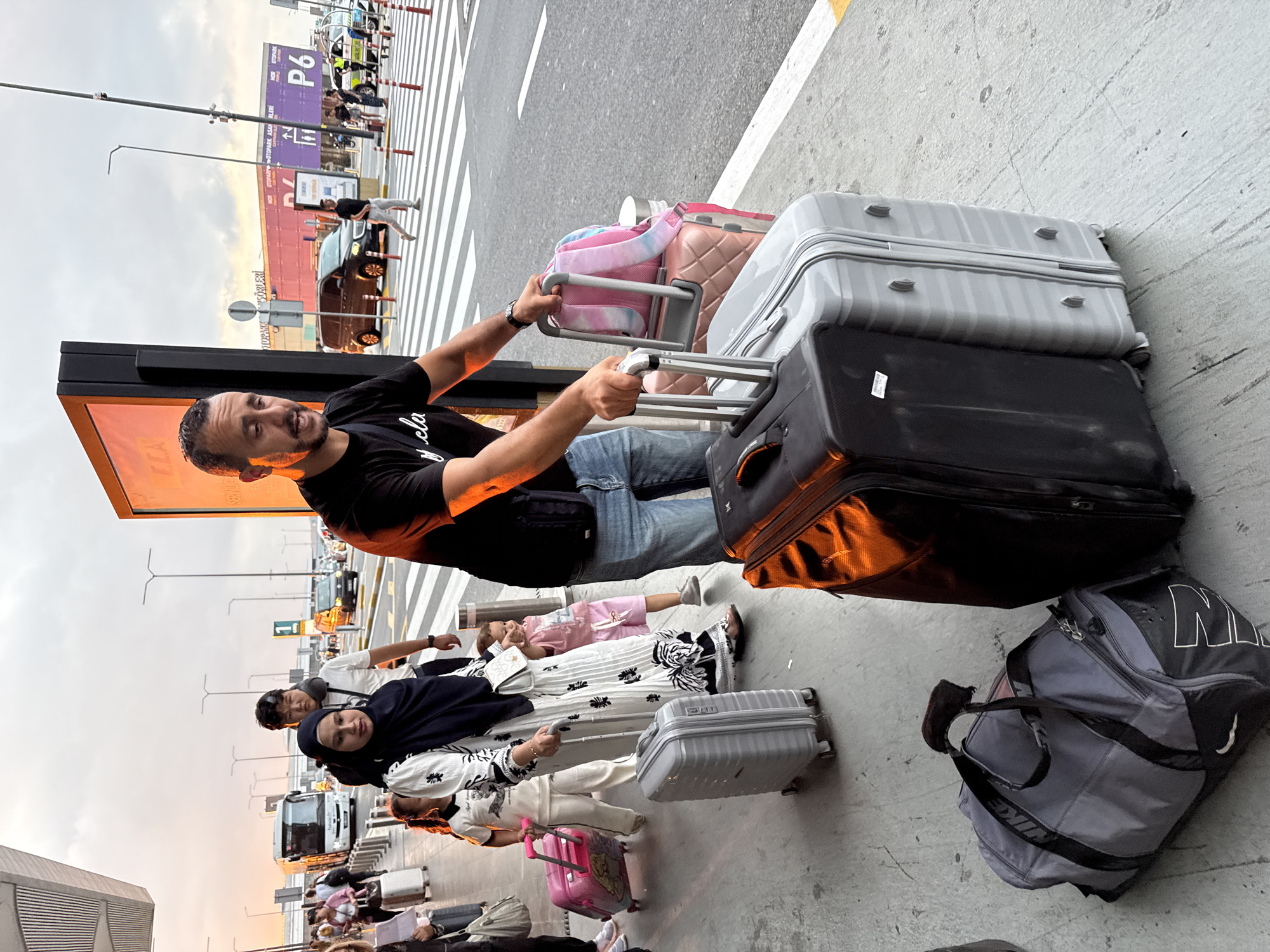 The image captures a family with luggage at an airport terminal in Arnavutköy, Turkey. A man in a black t-shirt and blue jeans is holding the handle of a light gray suitcase. Beside him, a pink suitcase is stacked with a colorful backpack. A woman in a black hijab and a white dress with black floral patterns is walking with a gray suitcase. Two young girls are with her; one in a white outfit and the other in pink shorts and a t-shirt. A dark gray duffel bag rests on the ground in the foreground. In the background, a white bus, cars, and airport buildings are visible under a cloudy sky during what appears to be sunset. The scene is set on a paved walkway in front of the airport.