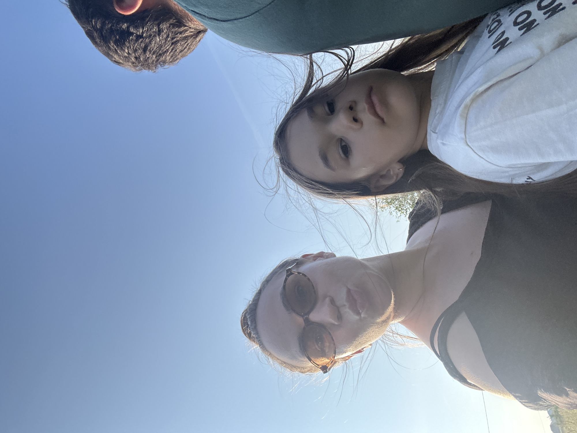 In Kharnavy, Russia, a low-angle, wide-shot selfie captures two individuals against a bright, clear sky. On the left, a woman wearing sunglasses and a black off-the-shoulder top puckers her lips, her hair blowing in the wind. To her right, a young girl in a white t-shirt with text visible looks forward, her hair also windswept. The back of another person, wearing a green shirt, is visible on the far right of the frame. The background suggests an outdoor setting, possibly rural, with a hint of trees and a structure in the distance on the left. The lighting indicates it is daytime, likely late afternoon due to the warm glow on the subjects.