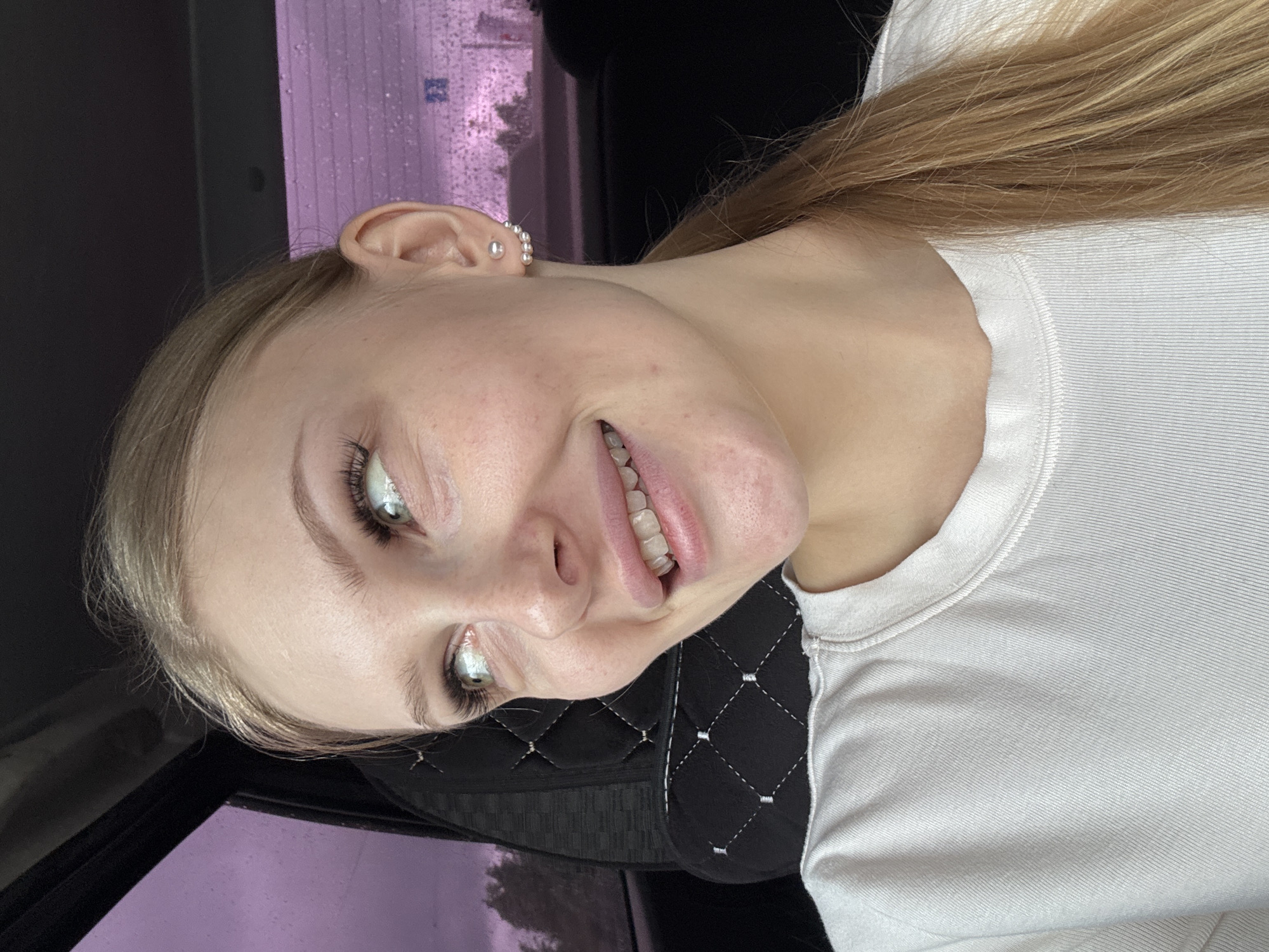 A young, blonde woman is seated in a car. She is turned to her right, smiling slightly, with her face directed toward the camera. She is wearing a white, ribbed, short-sleeved shirt. Her left ear is visible, adorned with several small, silver, rounded earrings. The car interior is visible, including a black seat with white stitching. Outside the car window, raindrops can be seen on the glass, and the cityscape of what is presumed to be Perm, Russia, is slightly obscured. The specific location within Perm cannot be determined.