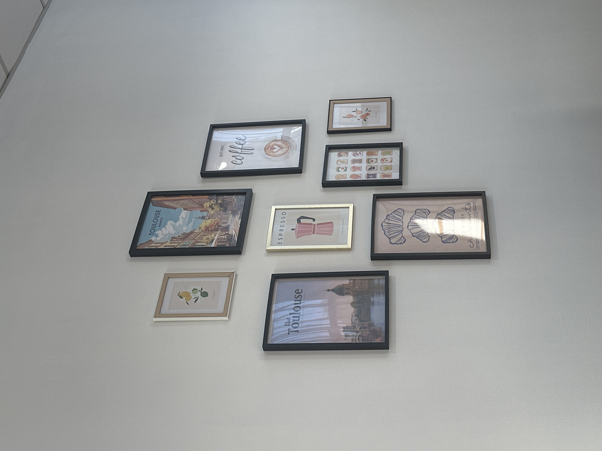 The image shows a collection of framed prints on a white wall. The prints vary in size and style, depicting coffee-related themes, pastries, and cityscapes, some possibly representing Toulouse, France. The framing is consistent across several prints, predominantly black, with one gold frame. The arrangement of the prints on the wall appears somewhat random or informal. The image does not reveal a specific location in Ramonville-Saint-Agne, France, or depict people or activities.